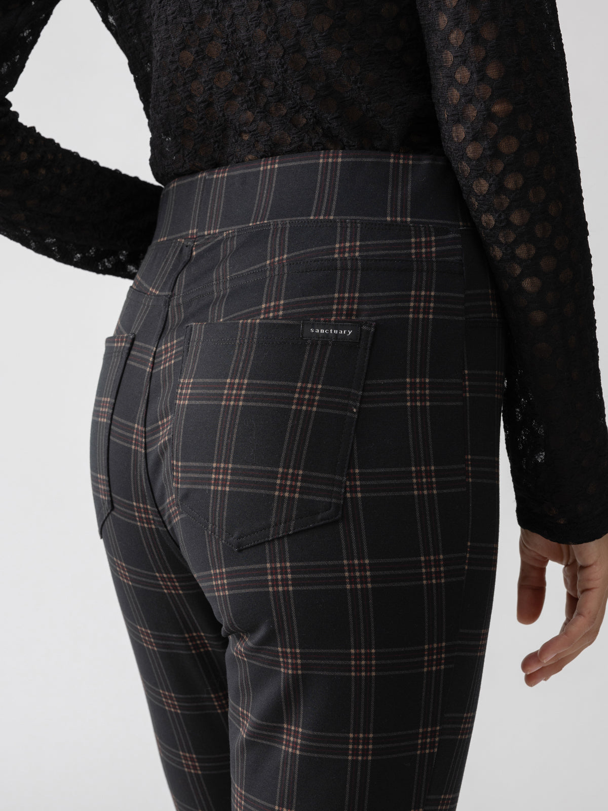 A person wearing Sanctuary Clothing's 'Runway Legging Garnet Plaid' and a black lacy top is shown from the back in this image. The leggings feature a plaid pattern of horizontal and vertical lines forming squares and a small label on the pocket. The person's arm is slightly visible on the right side.