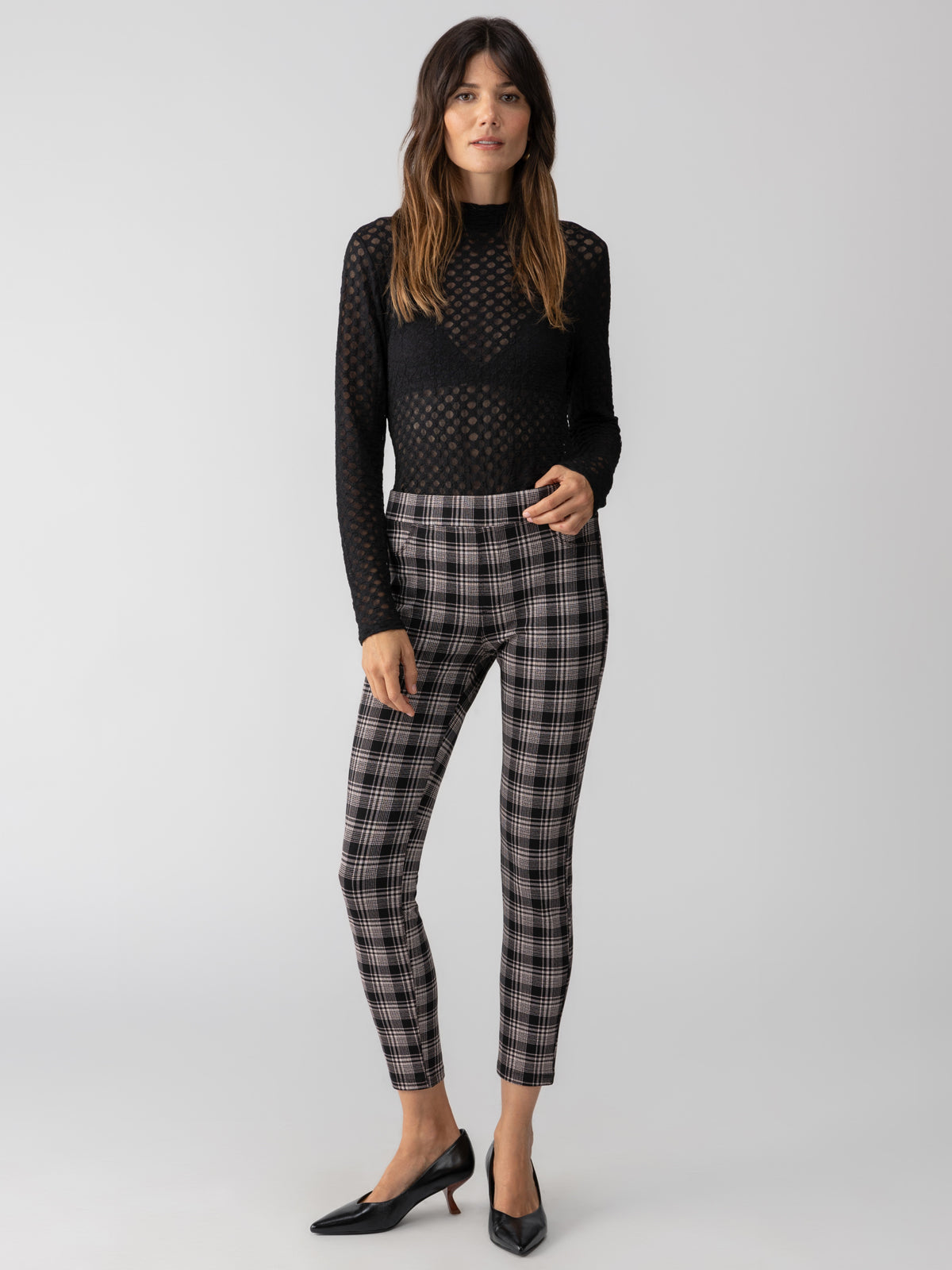 A woman stands against a plain background wearing a black, long-sleeve, see-through top over a black bra and the Runway Legging Almond Plaid by Sanctuary Clothing. She pairs her outfit with black pointed-toe heels. Her long hair is loose around her shoulders, and she has a neutral expression.