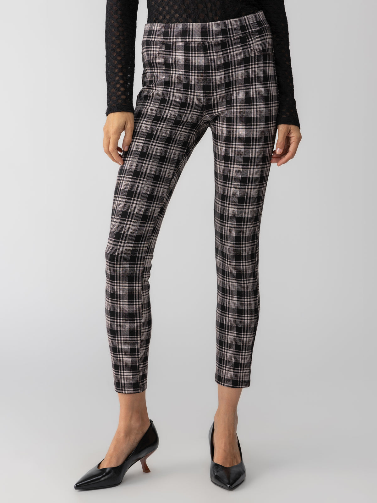 A person is wearing Sanctuary Clothing's Runway Legging Almond Plaid paired with a black long-sleeve top. The outfit is completed with black pointed-toe kitten heels. The background is plain and neutral, emphasizing the clothing and footwear.