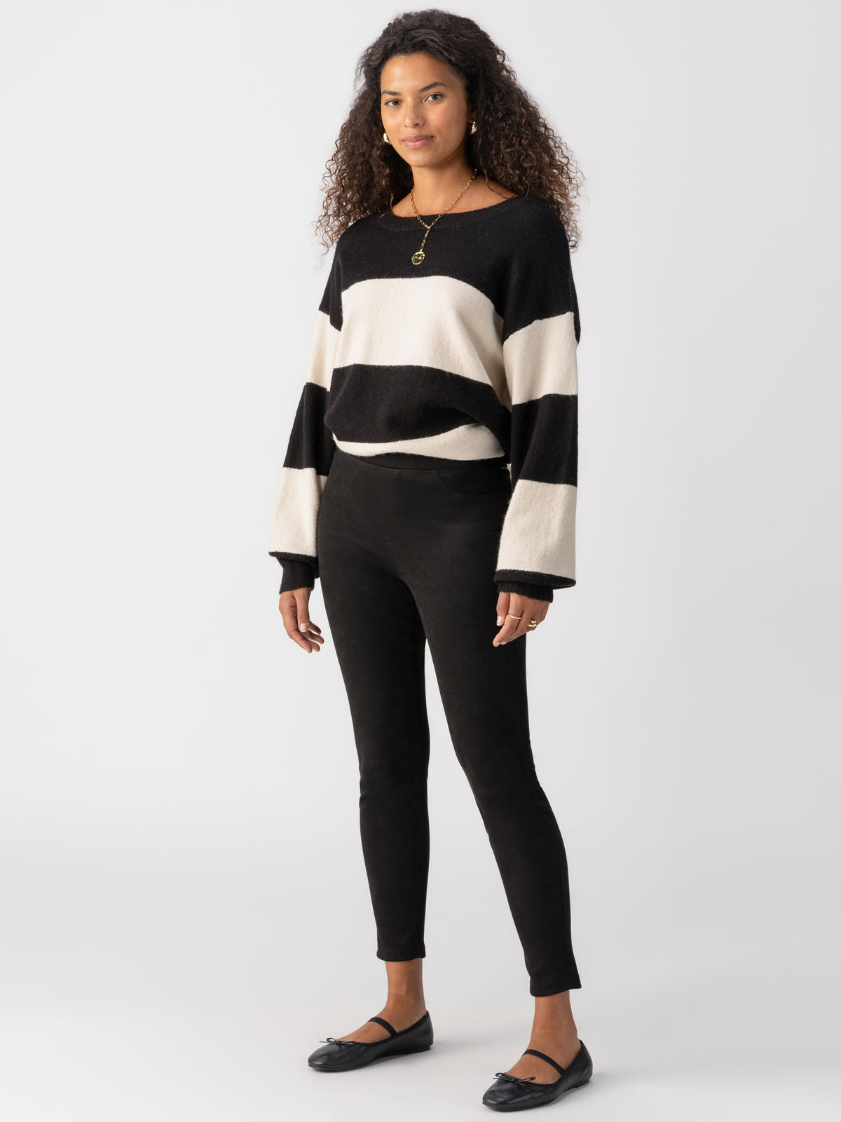 A woman with curly hair stands against a plain backdrop wearing a black and white striped sweater with wide sleeves, Sanctuary Clothing's Runway Legging Faux Suede Black, and black flats. She has a pendant necklace and a confident expression.