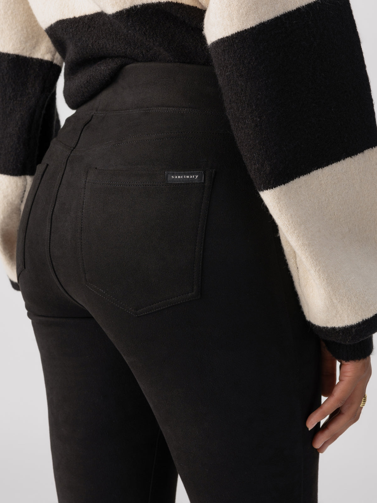 A person wearing a black and white striped sweater and Sanctuary Clothing's Runway Legging Faux Suede Black is shown from the back. The leggings have back pockets with a small label visible on one of the pockets. The background is plain and gray.