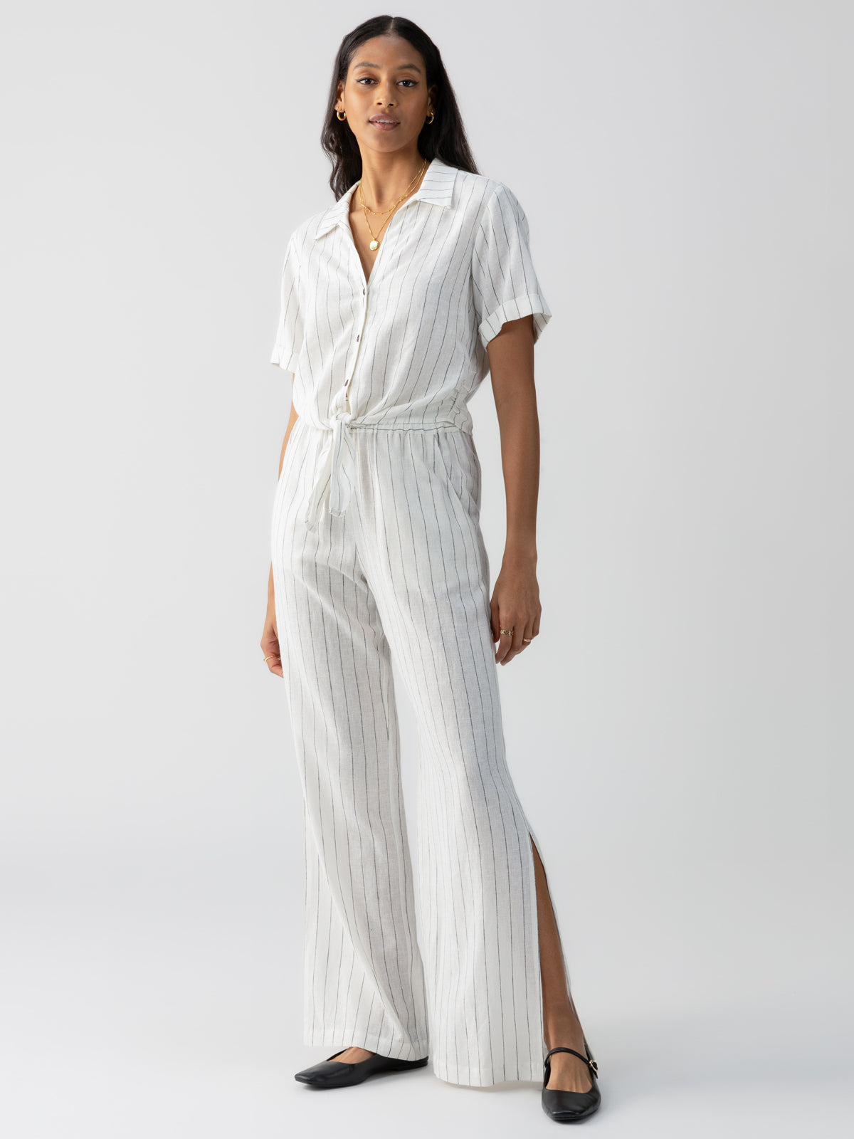 A woman stands against a plain background, wearing the Ocean Front Pull On Pant Biarritz Stripe from Sanctuary Clothing. The pants feature a white base with vertical stripes and have a relaxed, wide-leg fit. She pairs her outfit with black flats and gold hoop earrings.