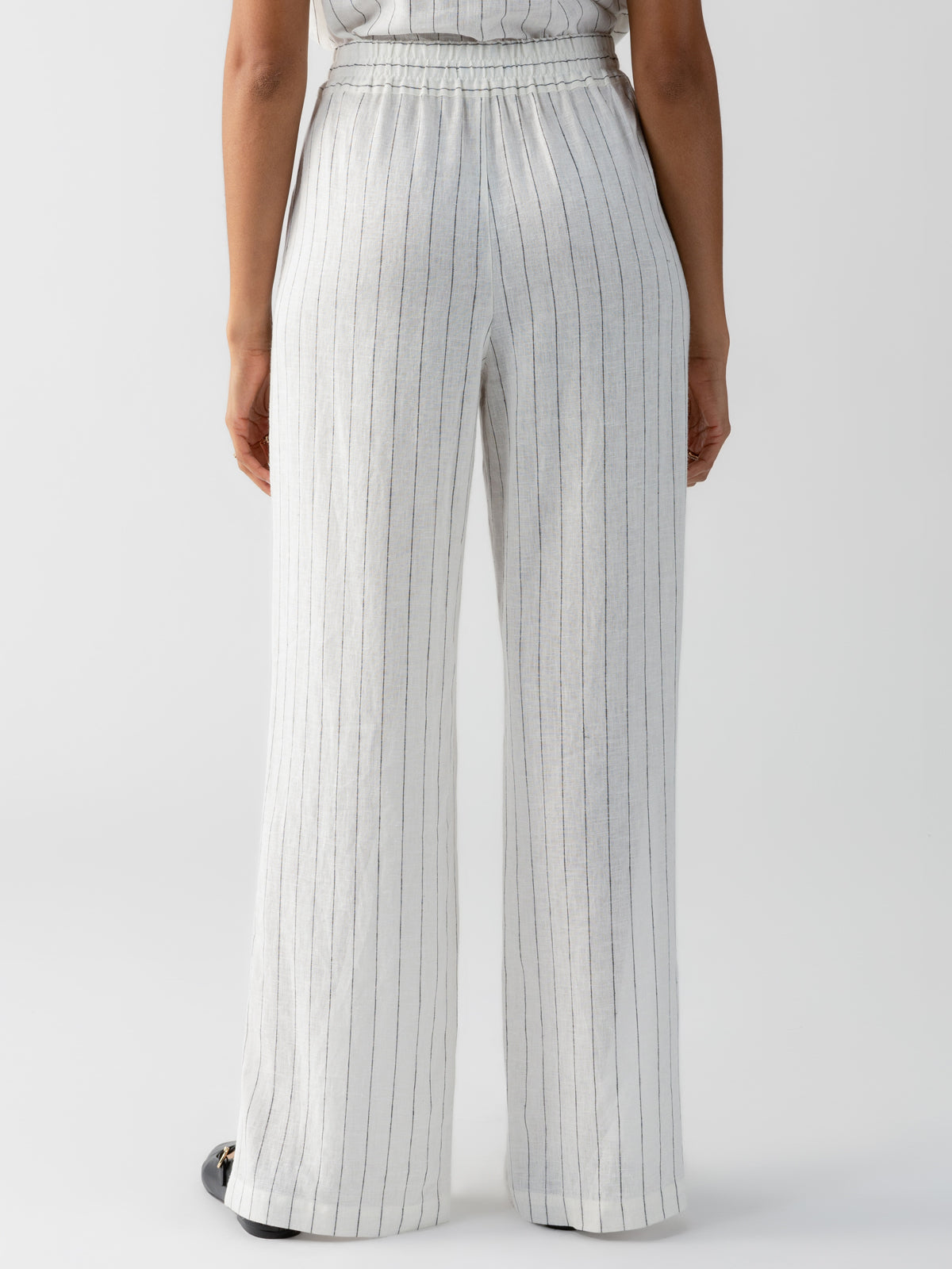 A person is standing facing away, wearing the Ocean Front Pull On Pant Biarritz Stripe from Sanctuary Clothing. The high-waisted, white pinstriped trousers feature a loose, straight fit and an elastic waistband. The background is plain and white.