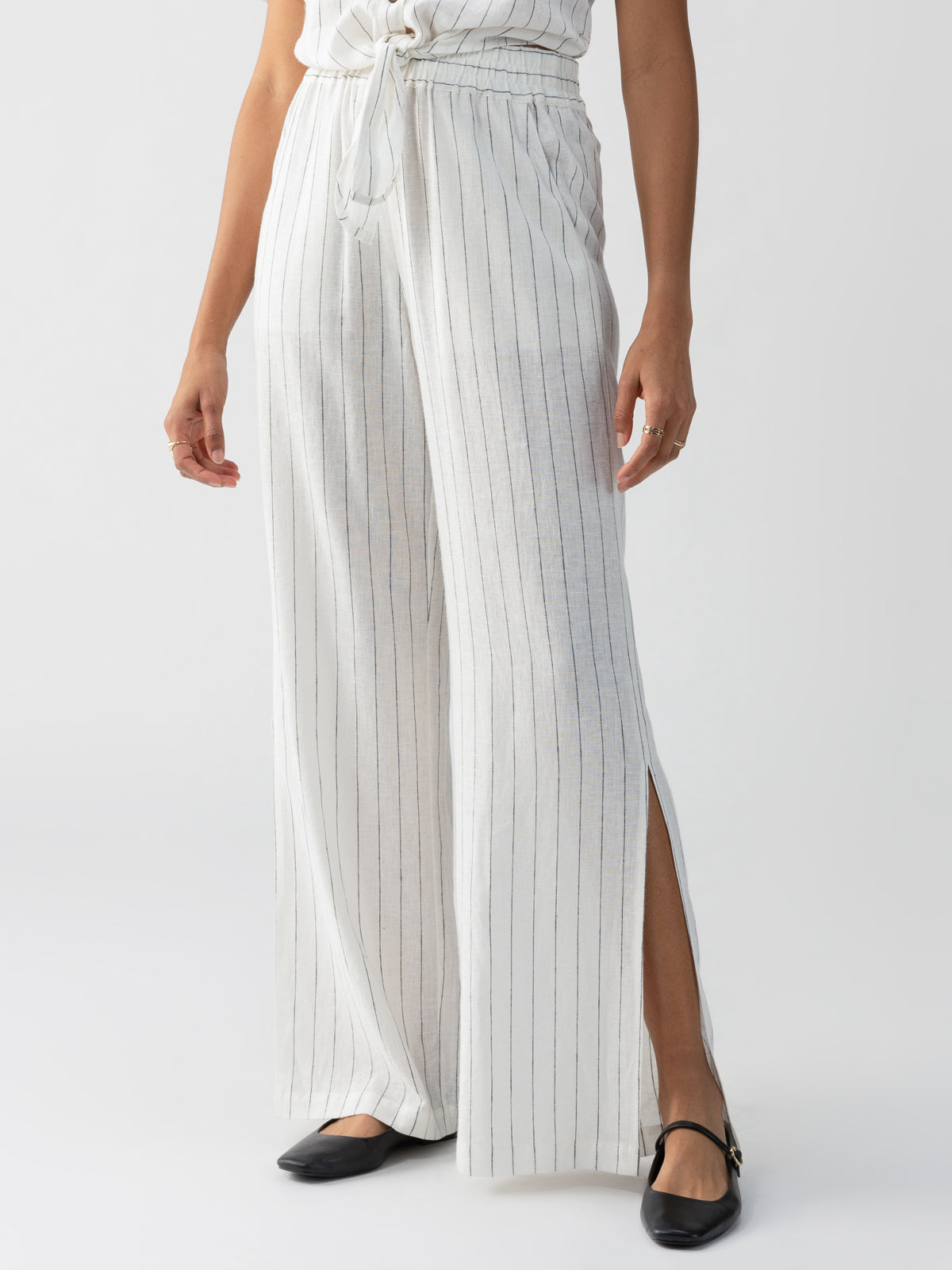 A person is wearing the Ocean Front Pull On Pant Biarritz Stripe by Sanctuary Clothing, which are high-waisted, white linen pants with thin vertical black stripes and side slits. The top part of their body is not visible. They are also wearing black flats with a strap across the top of the foot.