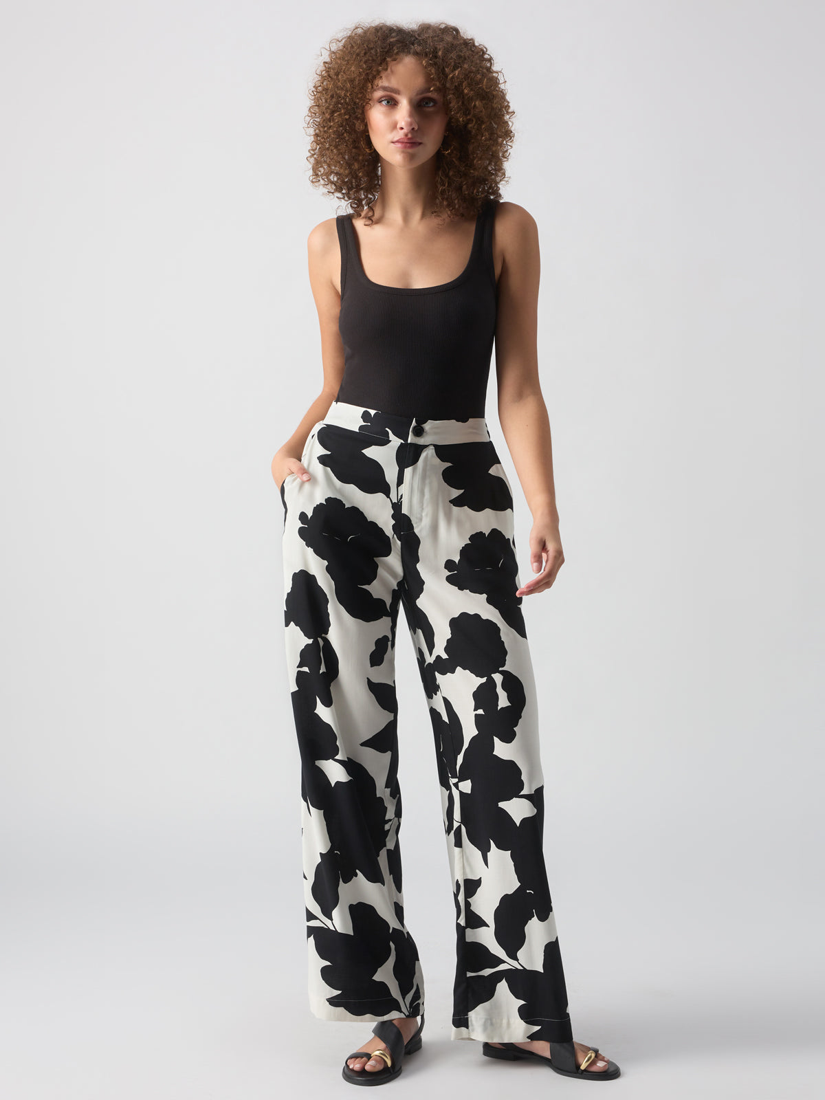 A woman with curly hair is wearing a black sleeveless top and Sanctuary Clothing's 'The Soft Trouser Spring Shadow,' a pair of high-waisted, wide-leg pants featuring a bold black and white floral pattern. She stands against a white background, looking forward with her hands in her pockets and wearing black sandals.