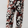 A person is shown in partial view, wearing Sanctuary Clothing's "The Soft Trouser" from the Mineral Inclusive Collection. These loose-fitting, wide-leg pants feature a bold abstract leaf print in black, white, and shades of reddish-brown. The individual has a hand in their pocket against a plain light gray background.