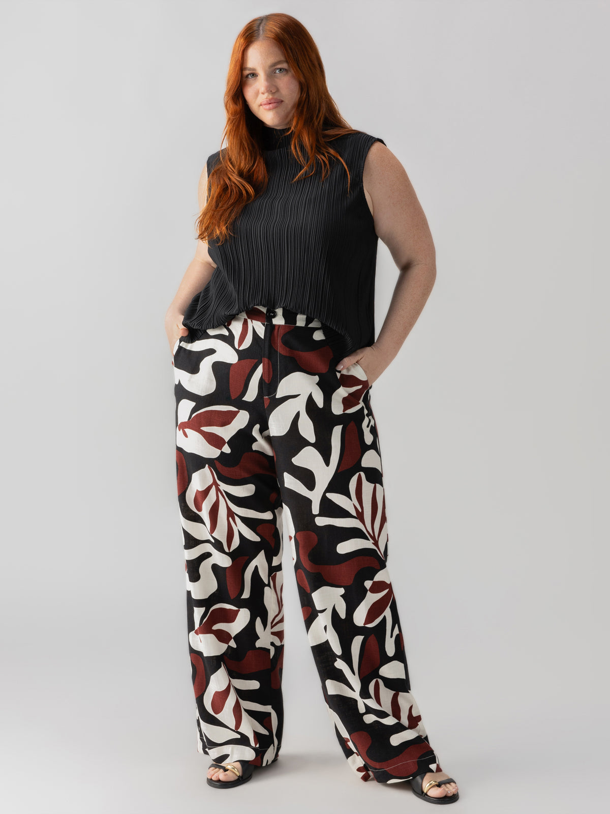 A woman with long red hair is standing against a plain background. She is wearing a sleeveless black top and The Soft Trouser Mineral Inclusive Collection by Sanctuary Clothing, which features a bold leaf pattern in black, white, and red. With her hands in her pockets, she exudes confidence.