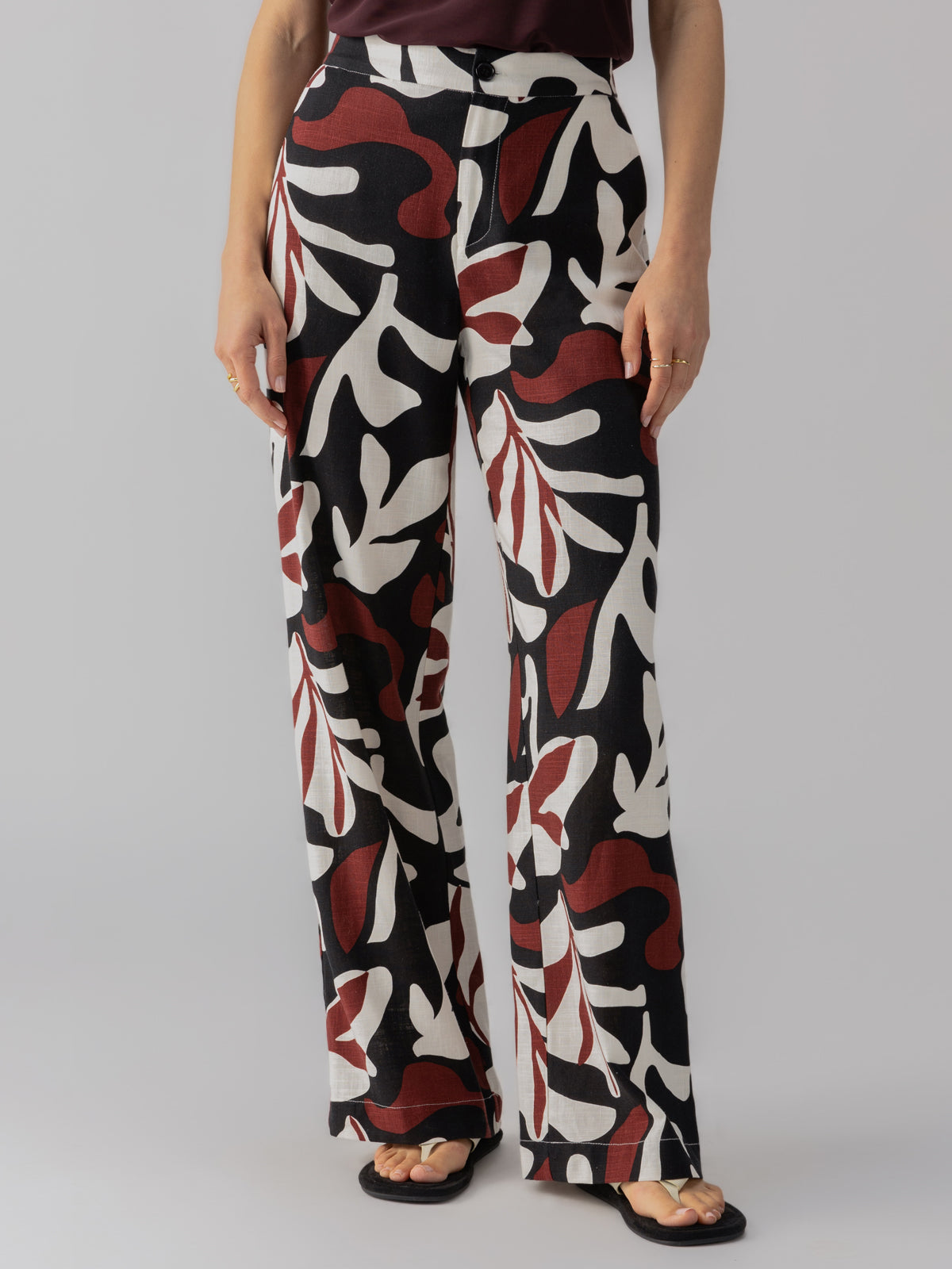 A person is wearing The Soft Trouser Mineral from Sanctuary Clothing, featuring a high-waisted, flowy fit and adorned with a black, white, and red abstract leaf pattern. The individual pairs the trousers with a dark top and black sandals. Only the lower half of the person is visible against a plain gray background.