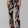 A person is wearing The Soft Trouser Mineral from Sanctuary Clothing, featuring a high-waisted, flowy fit and adorned with a black, white, and red abstract leaf pattern. The individual pairs the trousers with a dark top and black sandals. Only the lower half of the person is visible against a plain gray background.