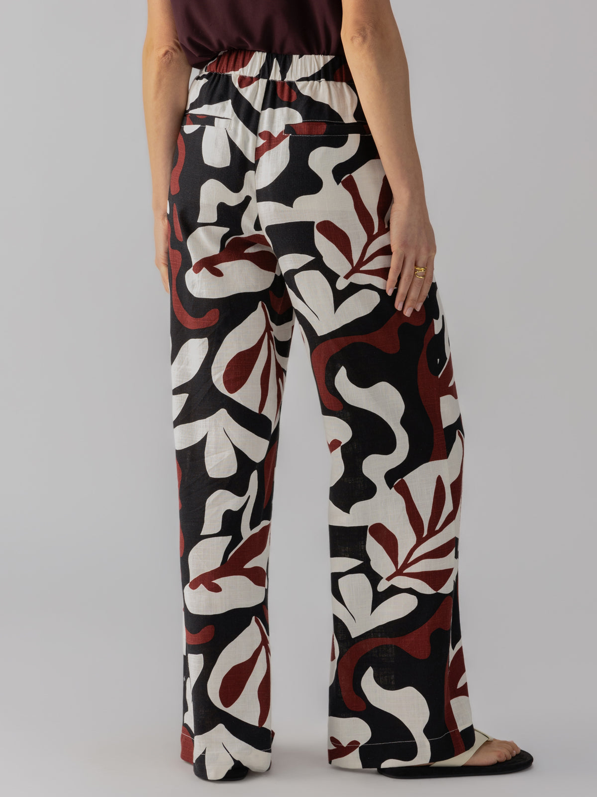 A person stands facing away, wearing The Soft Trouser Mineral by Sanctuary Clothing. These loose-fitting pants feature an abstract, bold pattern in black, white, and maroon. They include a high elastic waistband and wide legs. The person complements the look with a dark-colored top and adorns a ring on their left hand.