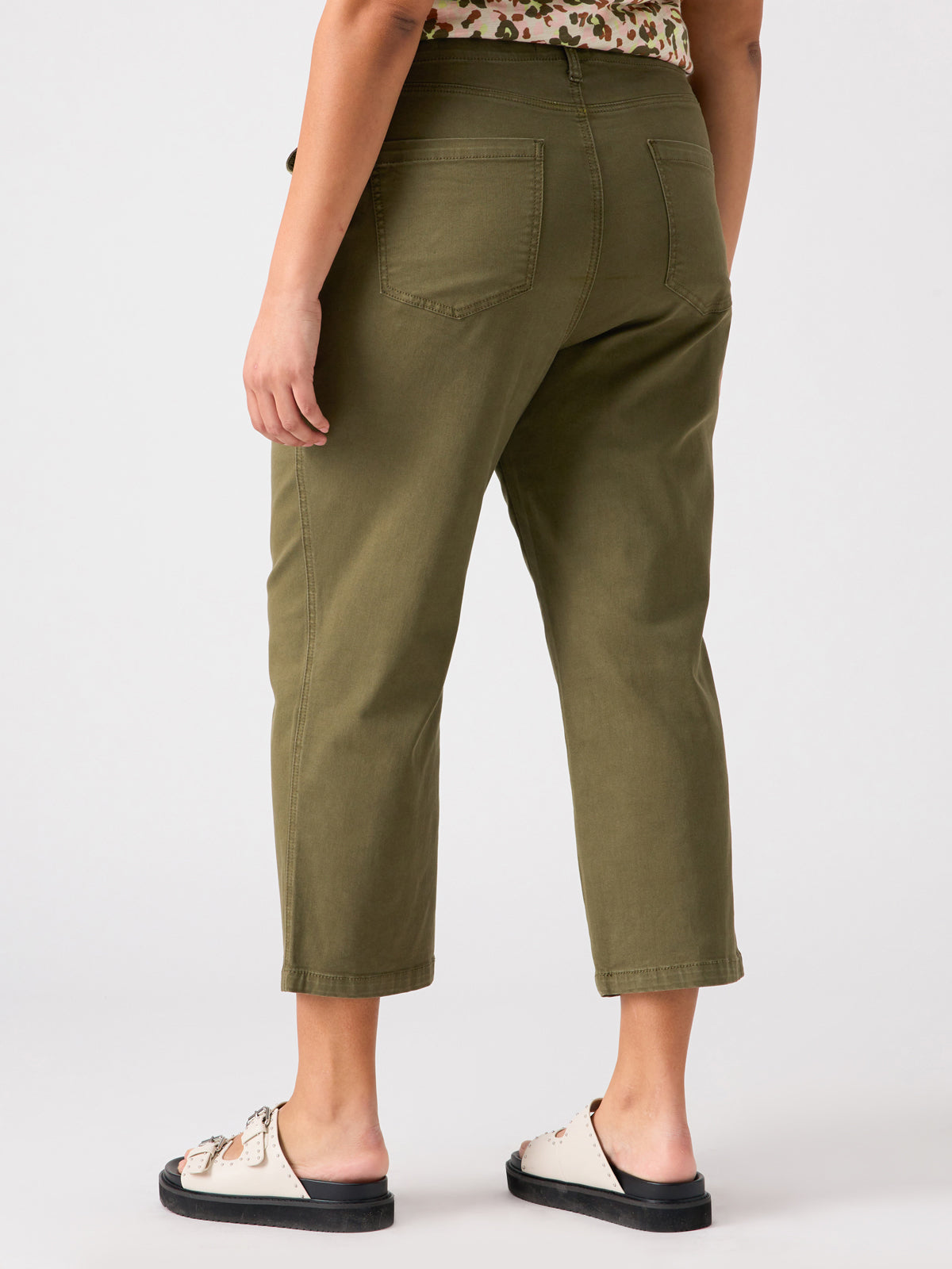 A person is wearing the Vacation Crop Burnt Olive Inclusive Collection trousers from Sanctuary Clothing and cream sandals with black soles. The photo is taken from behind, showing the back pockets of the trousers. The person is also wearing a patterned top.