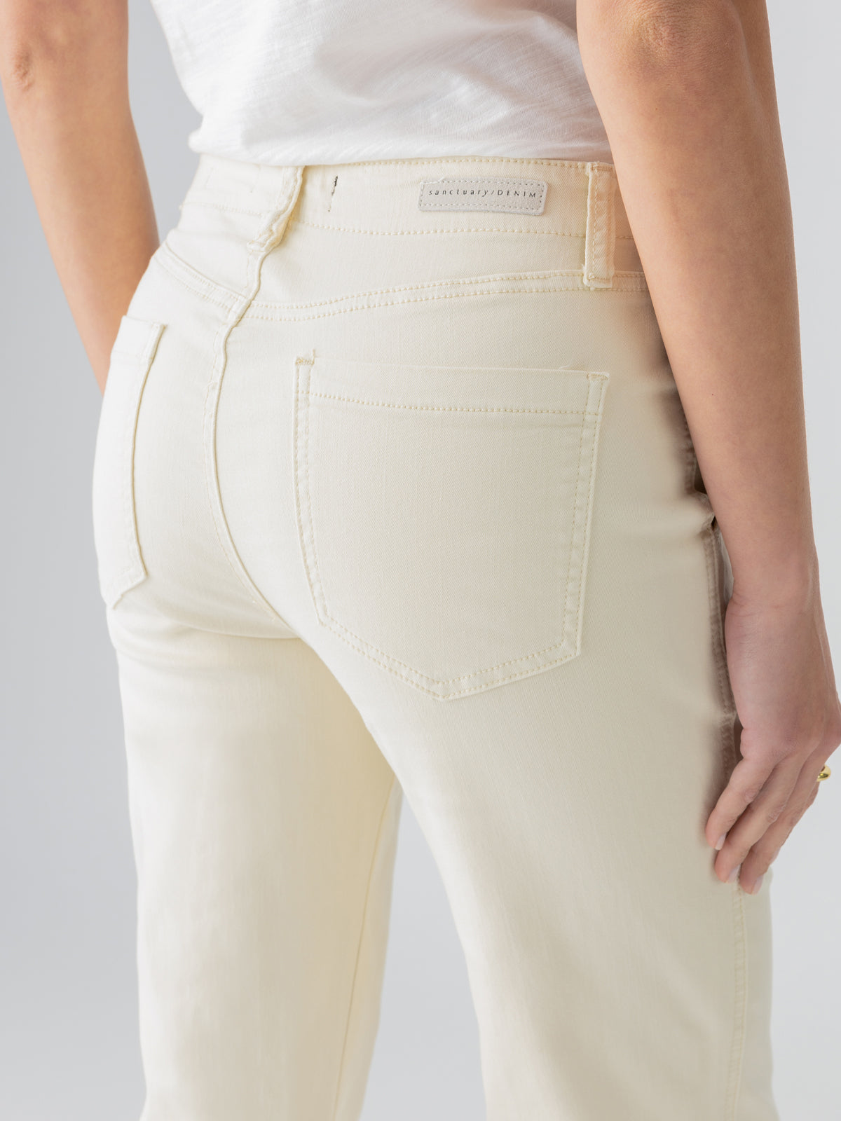 A person is wearing the "Vacation Crop Birch" high-waisted jeans from Sanctuary Clothing, featuring a cream color, two back pockets, and a stitched label near the waistband. They are paired with a white t-shirt, and only the lower back and hips are visible in the image.