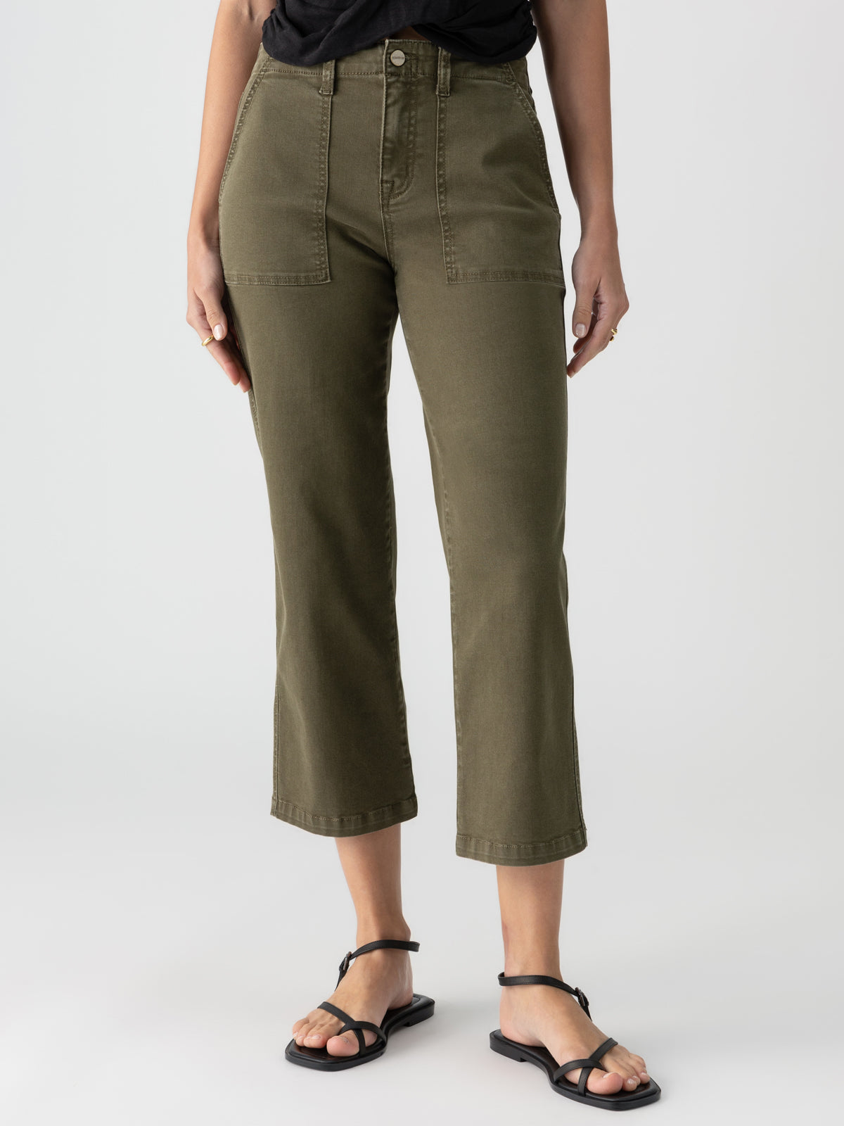 A person models the "Vacation Crop Burnt Olive" pants by Sanctuary Clothing, which feature large front pockets. They are pairing the olive green cropped pants with a black top and black strappy flat sandals. The background is a plain, light color, showing only their lower torso and legs.