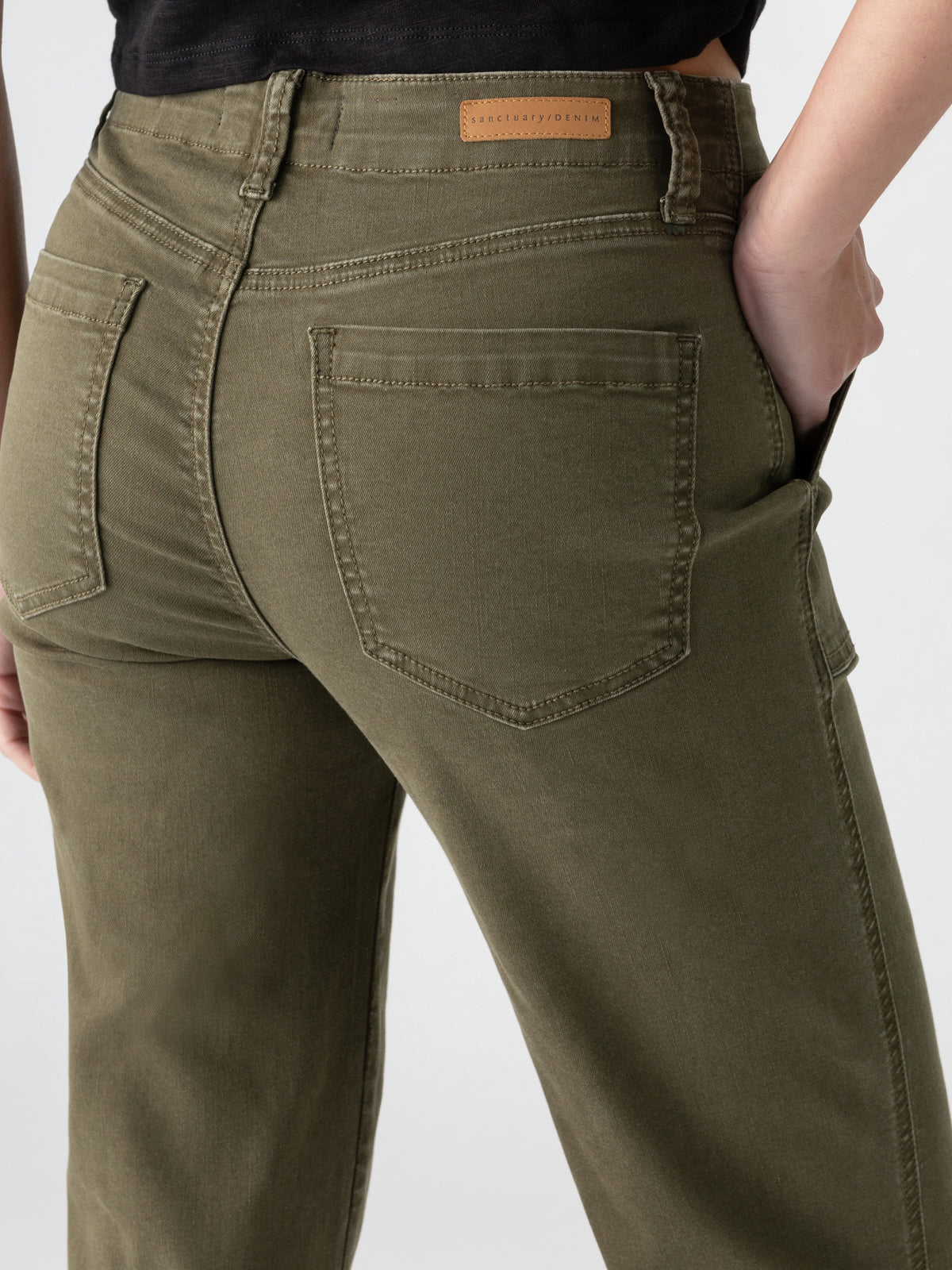 A person wearing the Vacation Crop Burnt Olive jeans from Sanctuary Clothing, featuring front pockets, stands with their hand in one pocket. The jeans boast visible stitching and a leather patch with text on the waistband at the back. The background is neutral.