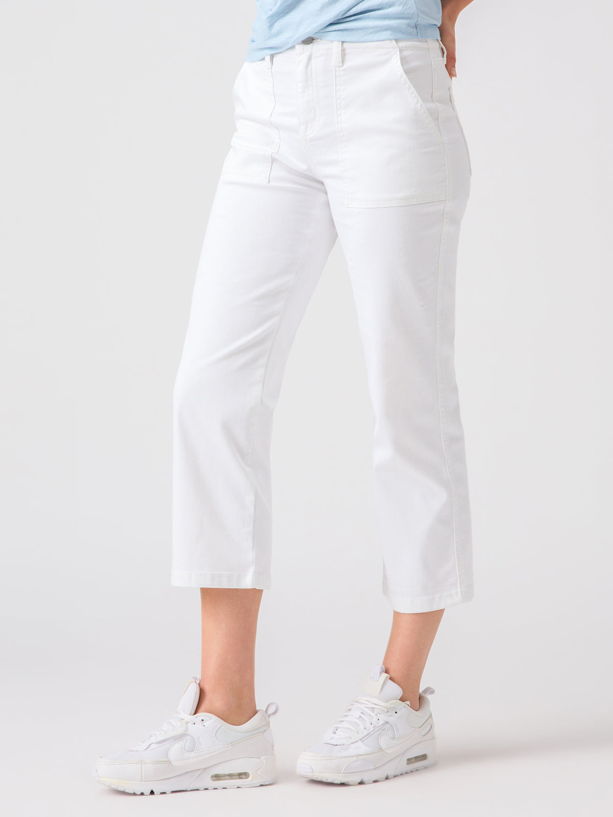 A person is sporting the Vacation Crop White pants by Sanctuary Clothing, paired with white sneakers. Their hands are tucked in the front pockets of the high-waisted cropped pants, and they stand against a plain light grey background. A light blue shirt is partially visible.