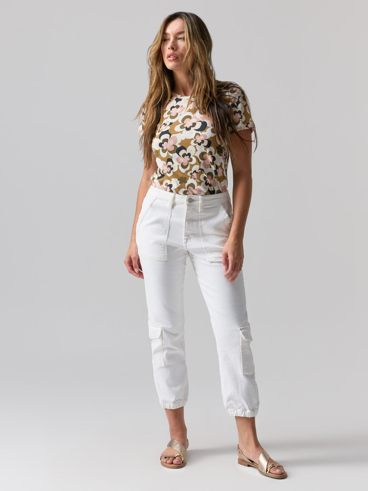 A woman with long, wavy hair stands against a plain background. She is wearing a floral-patterned t-shirt and the Brooklyn Cargo Pant Chalk from Sanctuary Clothing, featuring front pockets. She has on open-toed sandals and is posing with her hands by her sides, looking confidently ahead.