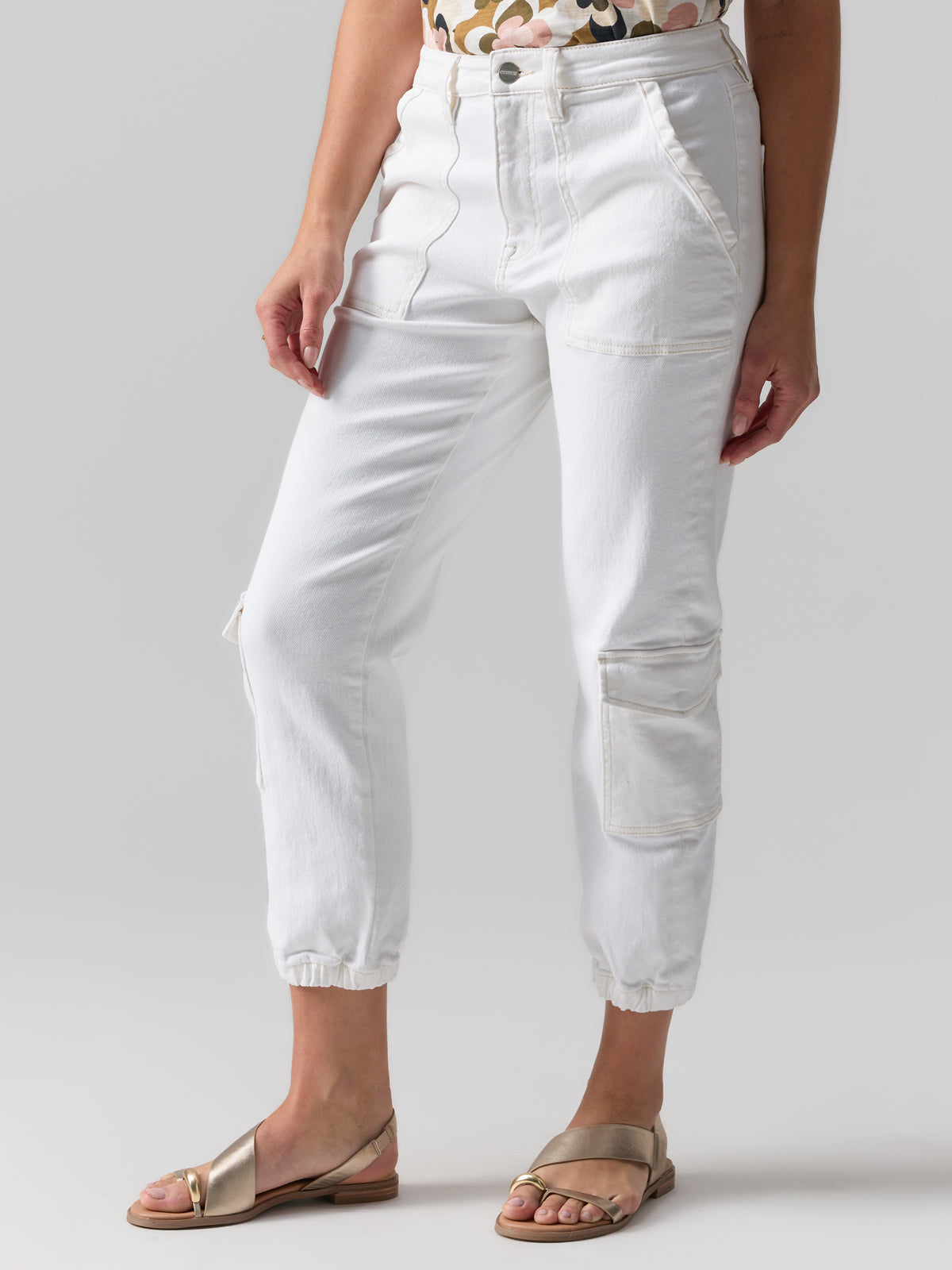 A person is wearing the Brooklyn Cargo Pant Chalk by Sanctuary Clothing, featuring large pockets and elastic cuffs, paired with metallic sandals. A sliver of a patterned top is visible against a plain, neutral background.