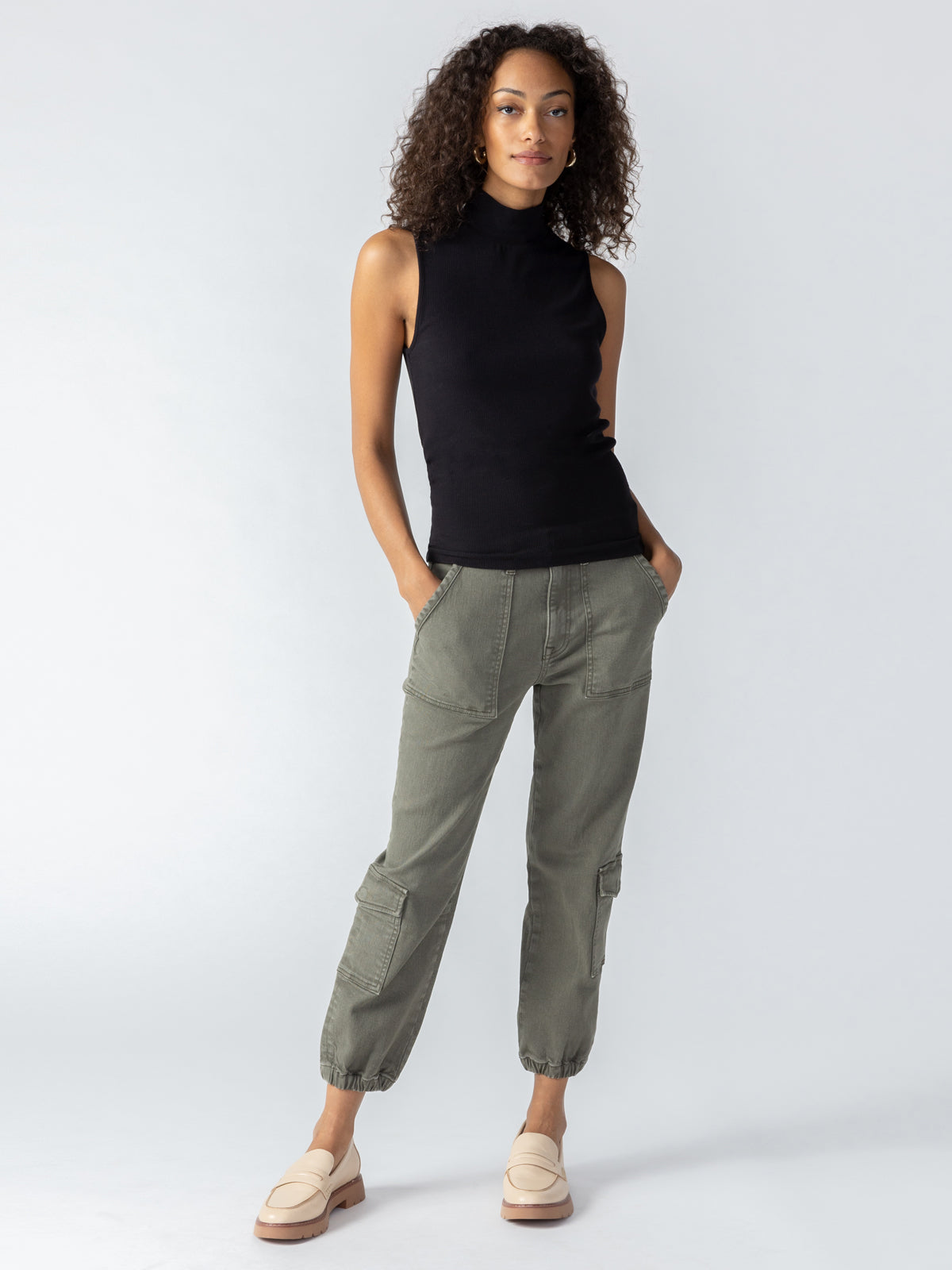 A person stands against a neutral background, wearing a sleeveless black turtleneck top, beige loafers, and the Brooklyn Cargo pants in Mossy Green from Sanctuary Clothing. Hands are in the pockets, and the individual has shoulder-length curly hair.