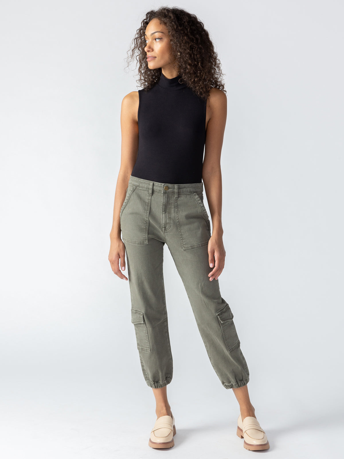 A woman with curly hair wears a sleeveless black top and the Brooklyn Cargo Mossy Green pants by Sanctuary Clothing, featuring a high-waisted design and elastic cuffs. Standing slightly to the right with her hands relaxed at her sides, she completes her casual outfit with white loafers.