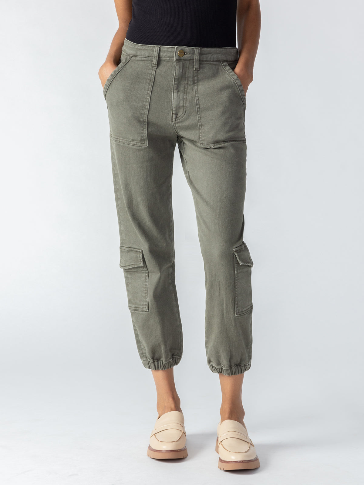 A person wearing the Sanctuary Clothing Brooklyn Cargo pants in Mossy Green, which feature elastic cuffs and multiple front and side pockets, along with beige loafers. Their hands are tucked into their pockets, and they are standing against a plain, light background.