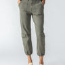 A person wearing the Sanctuary Clothing Brooklyn Cargo pants in Mossy Green, which feature elastic cuffs and multiple front and side pockets, along with beige loafers. Their hands are tucked into their pockets, and they are standing against a plain, light background.