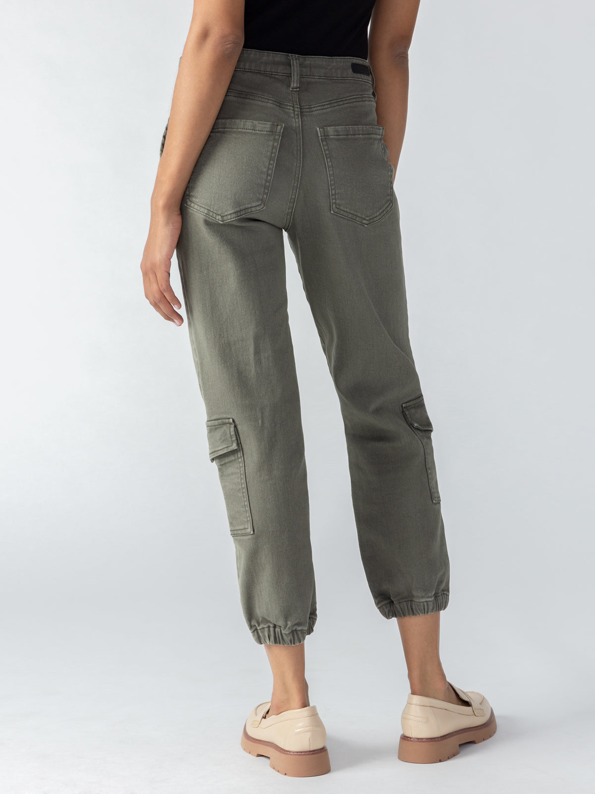 A person is standing with their back to the camera, wearing Brooklyn Cargo Mossy Green pants from Sanctuary Clothing, featuring pockets and elastic cuffs at the ankles. They are also wearing a black top and beige shoes. The background is a plain, light grey.