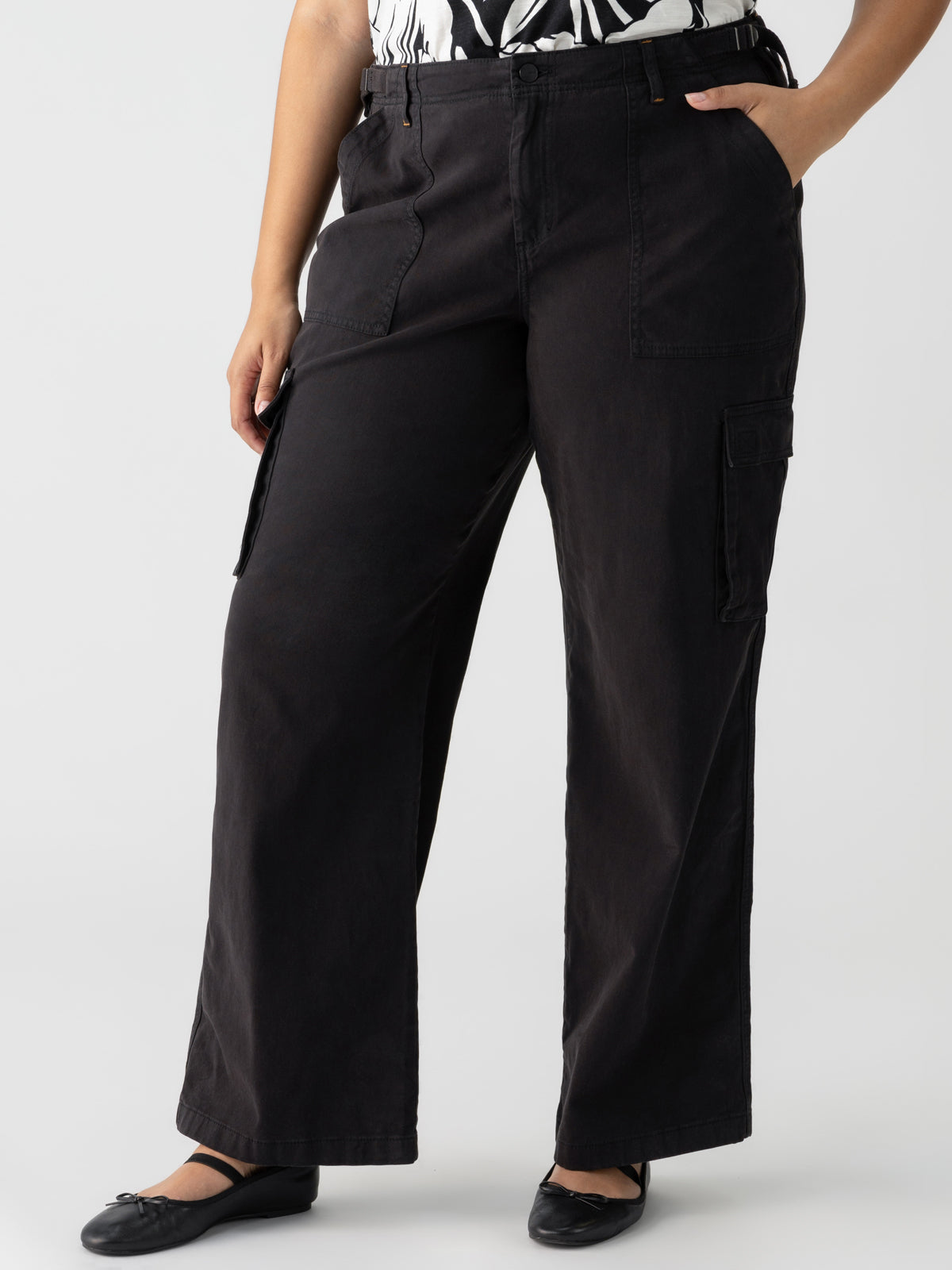 A person wearing Sanctuary Clothing's Reissue Cargo Pant Black from the Inclusive Collection, featuring a high-waisted fit with multiple pockets and a relaxed, wide-leg silhouette. They have paired it with a black-and-white printed top and are standing confidently with one hand on their hip and the other in their pocket. Completing the look, they are wearing black flats.