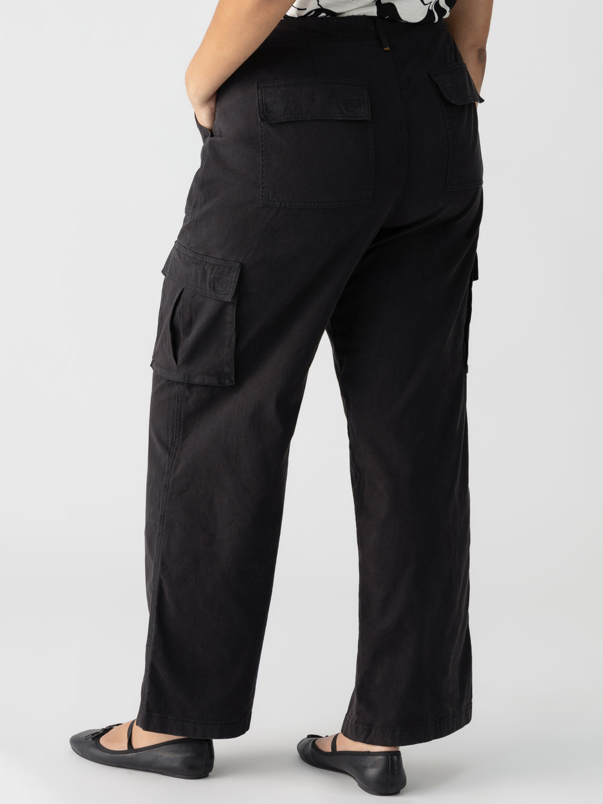 Rear view of an individual dressed in the Sanctuary Clothing Reissue Cargo Pant Black from the Inclusive Collection, featuring high-waisted design with flap pockets on both the back and sides. They are also wearing black flats against a plain white background.