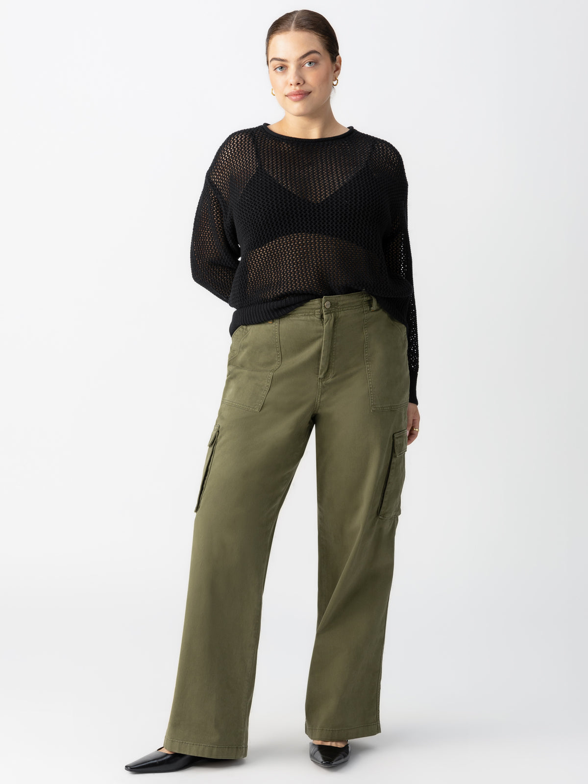 A person stands against a plain background wearing a black mesh long-sleeve top over a black tank top, paired with the Sanctuary Clothing Reissue Cargo Pant in Mossy Green from the Inclusive Collection and black shoes. Their hair is pulled back, and they have a neutral expression, with hands resting in the pockets of their pants.