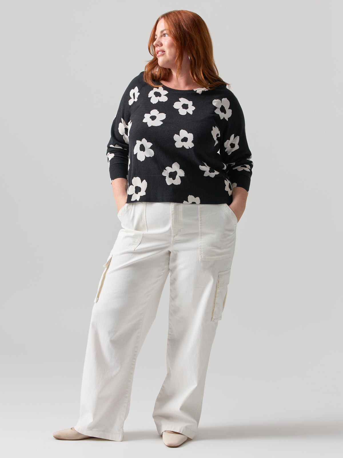 A woman with shoulder-length red hair stands against a plain background. She is dressed in a black sweater adorned with a large white flower pattern, white high-waisted wide-leg cargo pants from the Reissue Cargo Pant Powdered Sugar Inclusive Collection by Sanctuary Clothing, and beige shoes. Her hands are tucked into her pockets, and she gazes to the side with a slight smile.