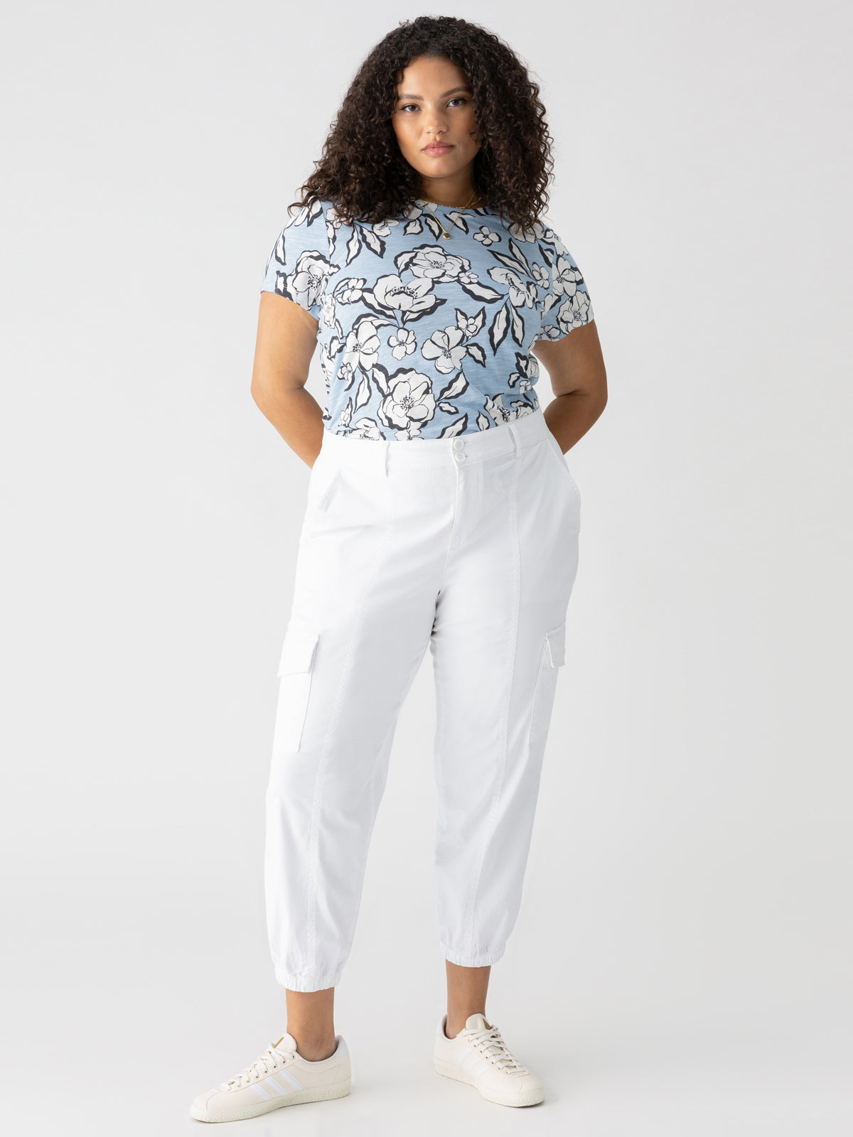 A person with curly hair is standing in a confident pose against a plain background. They are wearing a light blue floral-patterned shirt tucked into high-waisted Rebel Pant Eco Natural from the Inclusive Collection by Sanctuary Clothing, paired with white sneakers. Their hands are in their pockets, and they have a neutral expression.