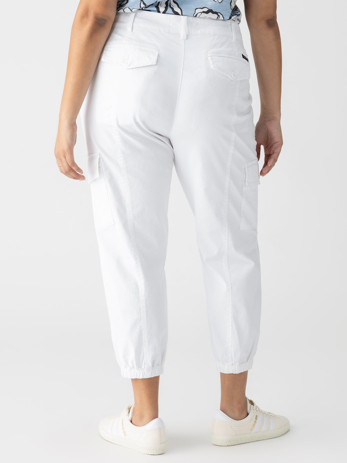 The image displays a person from the waist down, dressed in Sanctuary Clothing's Rebel Pant Eco Natural Inclusive Collection. The white cargo pants feature elasticated ankles and pockets on both the back and sides. The person is also wearing white sneakers adorned with beige stripes, along with a top that has a partially visible blue and white pattern.