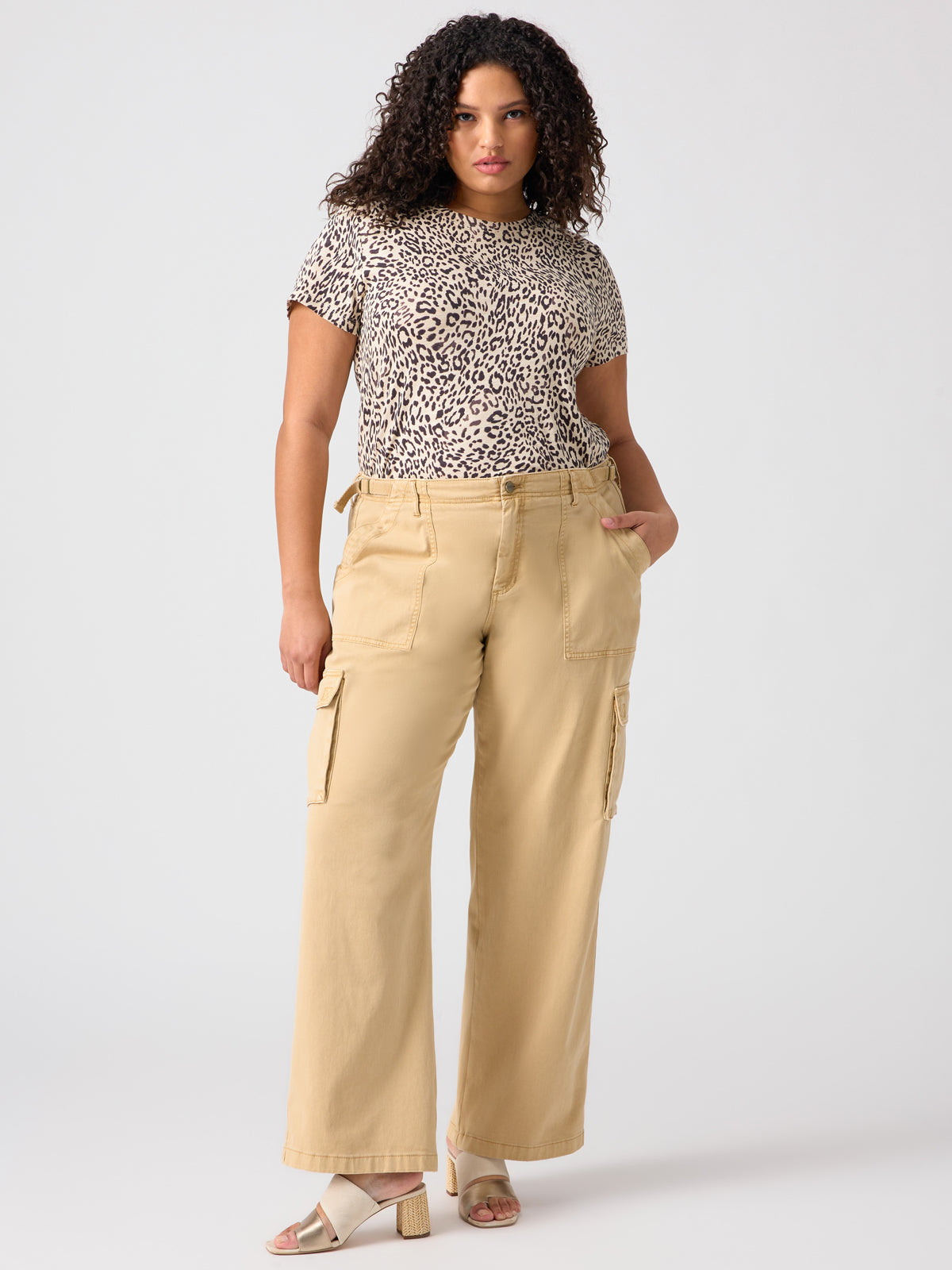 A woman with curly hair is wearing a leopard print T-shirt and Sanctuary Clothing's Reissue Cargo Pant in True Khaki from the Inclusive Collection. She is standing against a plain background with one hand in her pocket and the other hand resting by her side, while she completes her look with open-toe heeled sandals.