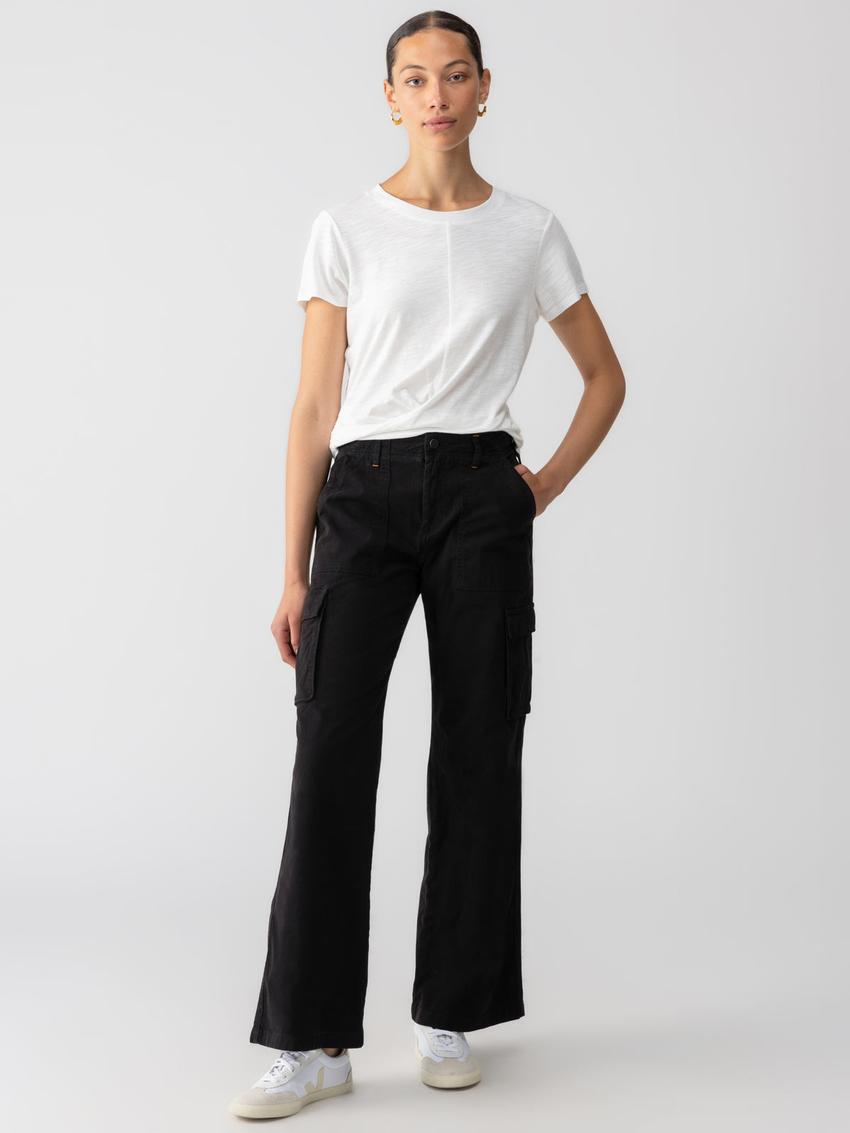 A person with a ponytail is standing against a plain background. They are wearing a white t-shirt, white sneakers, and the Reissue Cargo Black pants from Sanctuary Clothing. One hand is in their pocket, and the other rests by their side. They have gold hoop earrings.