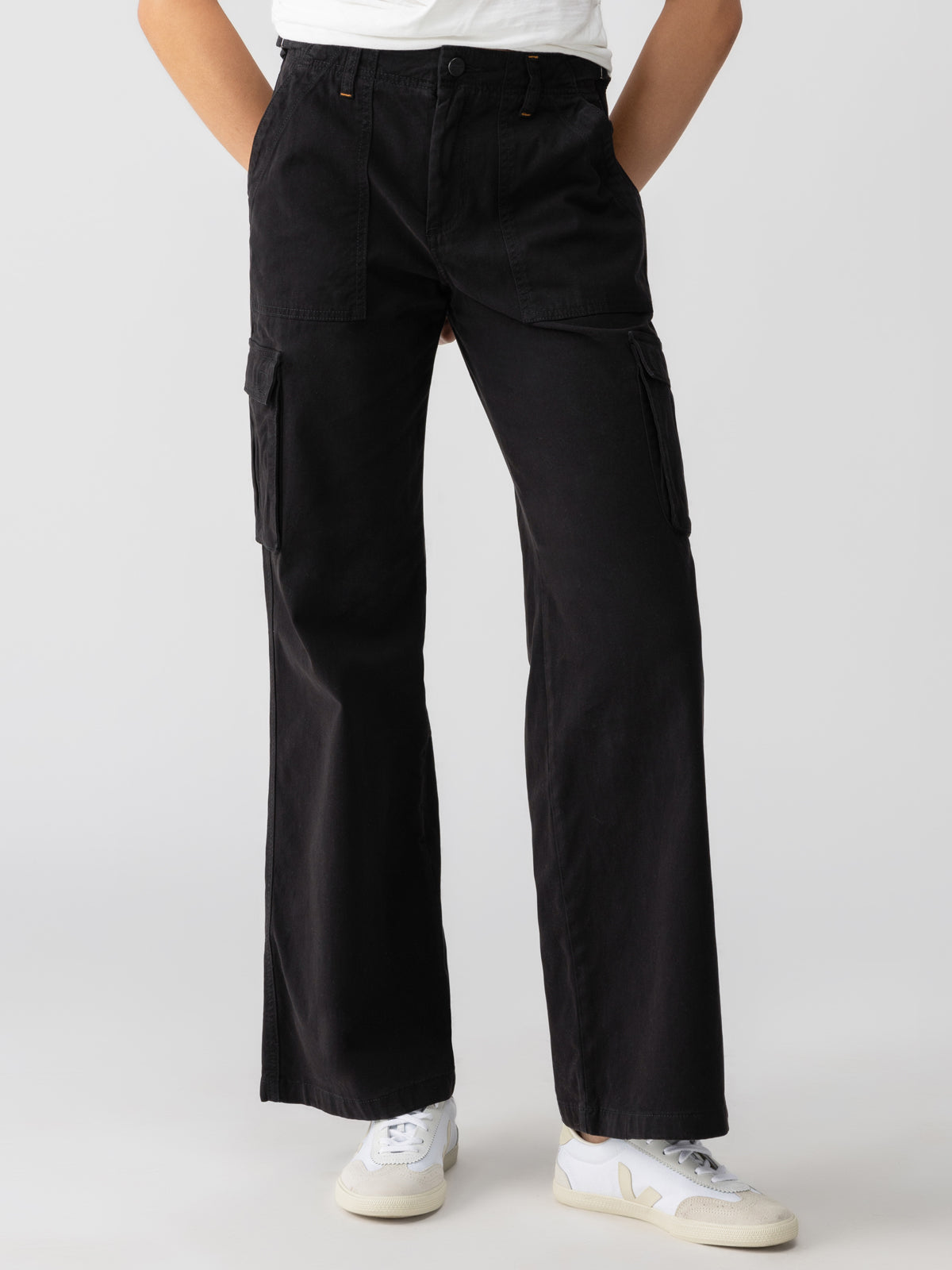 A person is wearing Sanctuary Clothing's Reissue Cargo Black wide-leg pants with large side pockets, paired with a white T-shirt. They are standing against a plain background, with hands in the pants' pockets. White sneakers with light-colored soles are visible on their feet.