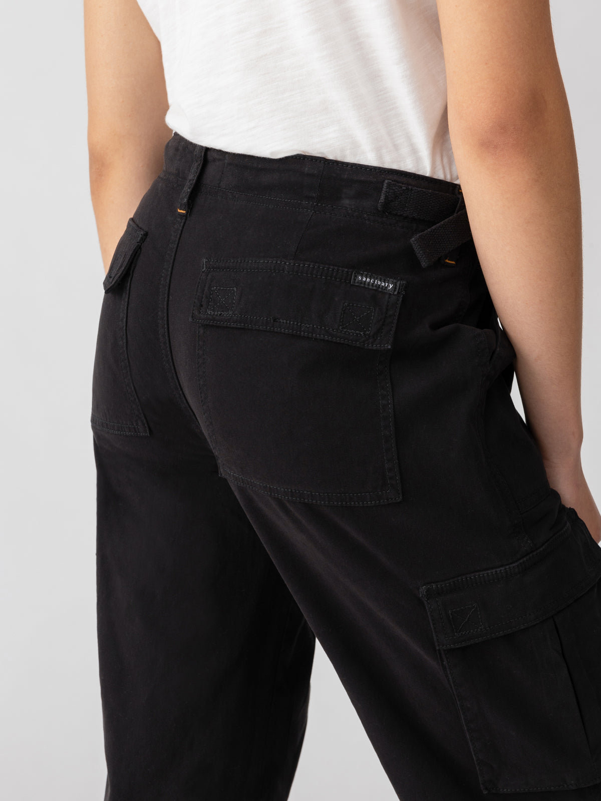 Close-up of a person wearing Sanctuary Clothing's Reissue Cargo Black pants, shown from the back. The pants feature large back pockets with flaps and button closures. The person is also wearing a white t-shirt. The background is neutral and minimalistic.