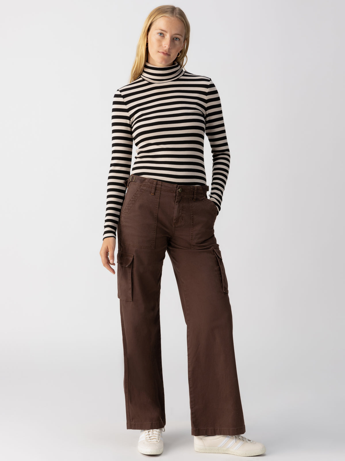 A person with long, light hair stands against a plain background, wearing a black and white striped long-sleeve turtleneck top paired with the Sanctuary Clothing Reissue Cargo Pants in Brown Sugar, and white sneakers. They have their hands in their pockets and are looking at the camera.