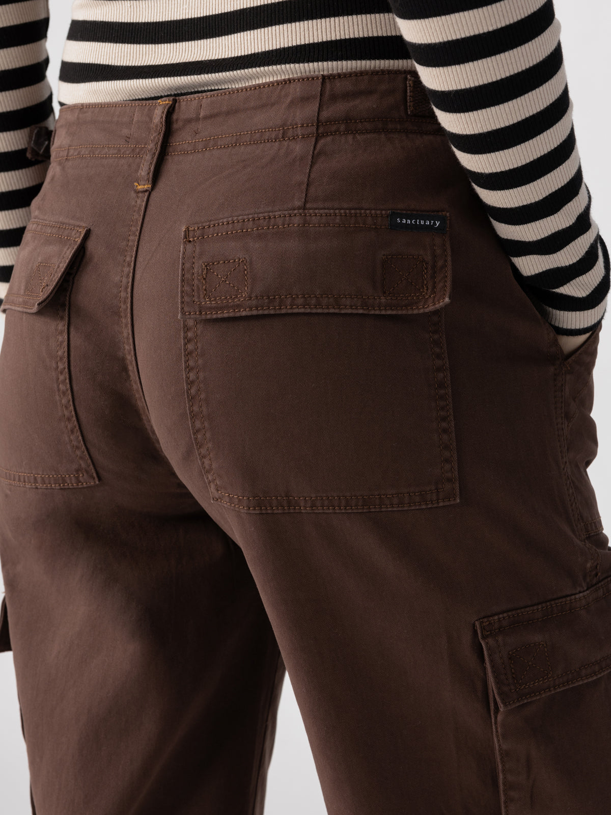 Close-up of a person wearing the Reissue Cargo Brown Sugar pants by Sanctuary Clothing, featuring large back pockets and paired with a striped long-sleeve shirt. A small black label is visible on one pocket. The durable fabric of these cargo pants makes them suitable for casual or workwear.