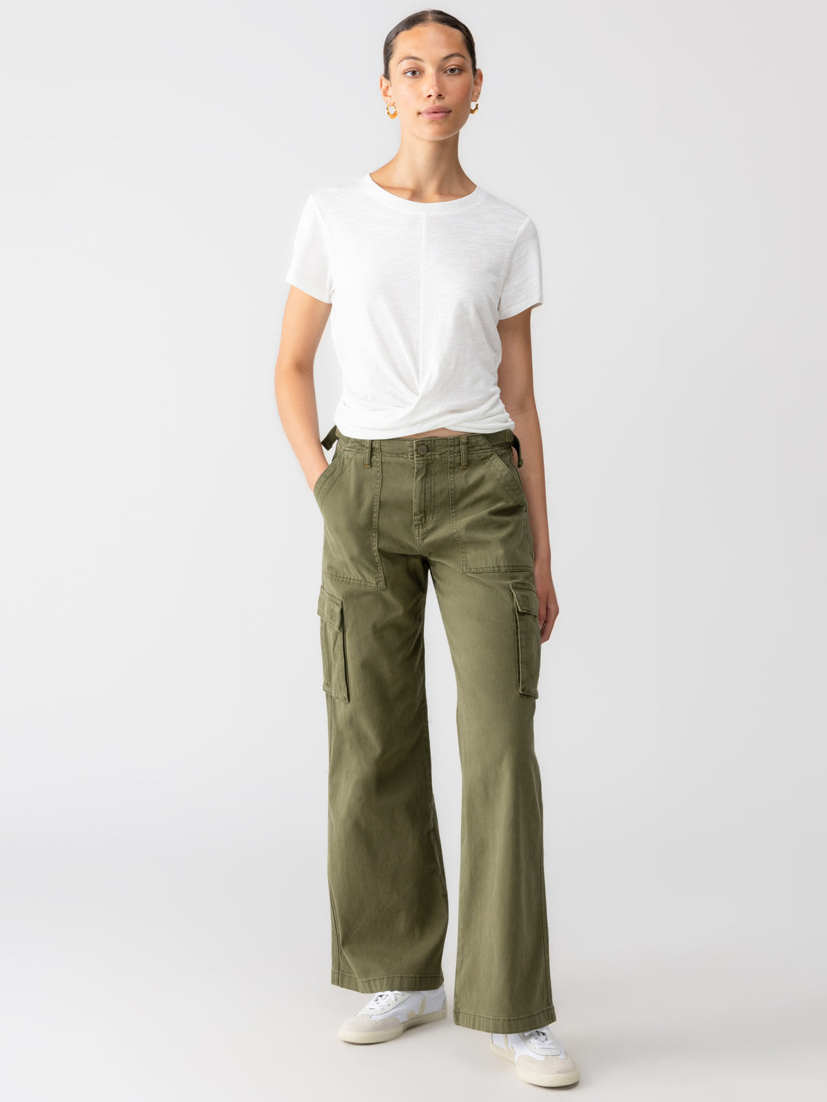 A person stands against a plain background wearing a white short-sleeve shirt and Sanctuary Clothing's Reissue Cargo Pant in Mossy Green with hands in pockets. They have short hair and are accessorizing with hoop earrings and white sneakers, creating a casual and relaxed look.