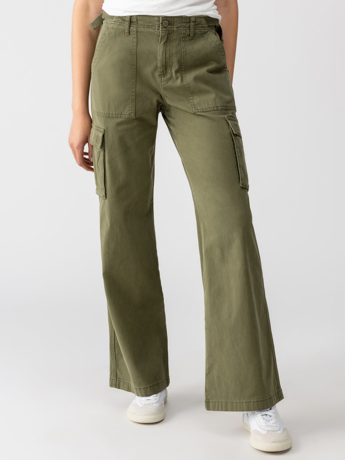 A person is wearing the Reissue Cargo Pant in Mossy Green from Sanctuary Clothing, which are high-waisted with multiple pockets, paired with a white T-shirt. They are also sporting white sneakers. The background is plain and light-colored.