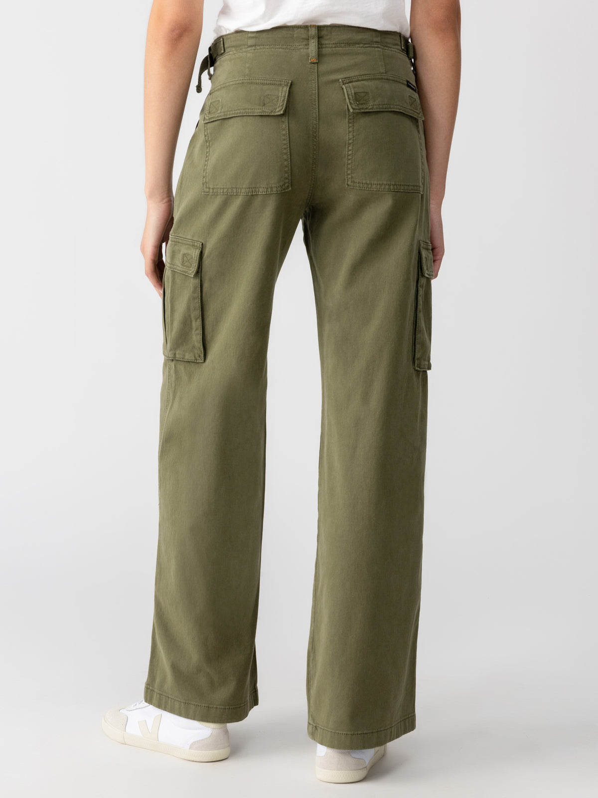 A person is standing with their back facing the camera, wearing Sanctuary Clothing's Reissue Cargo Pant in Mossy Green, featuring flap pockets on the back and sides. They are also wearing a white T-shirt and white sneakers with a gray logo. The background is light gray.