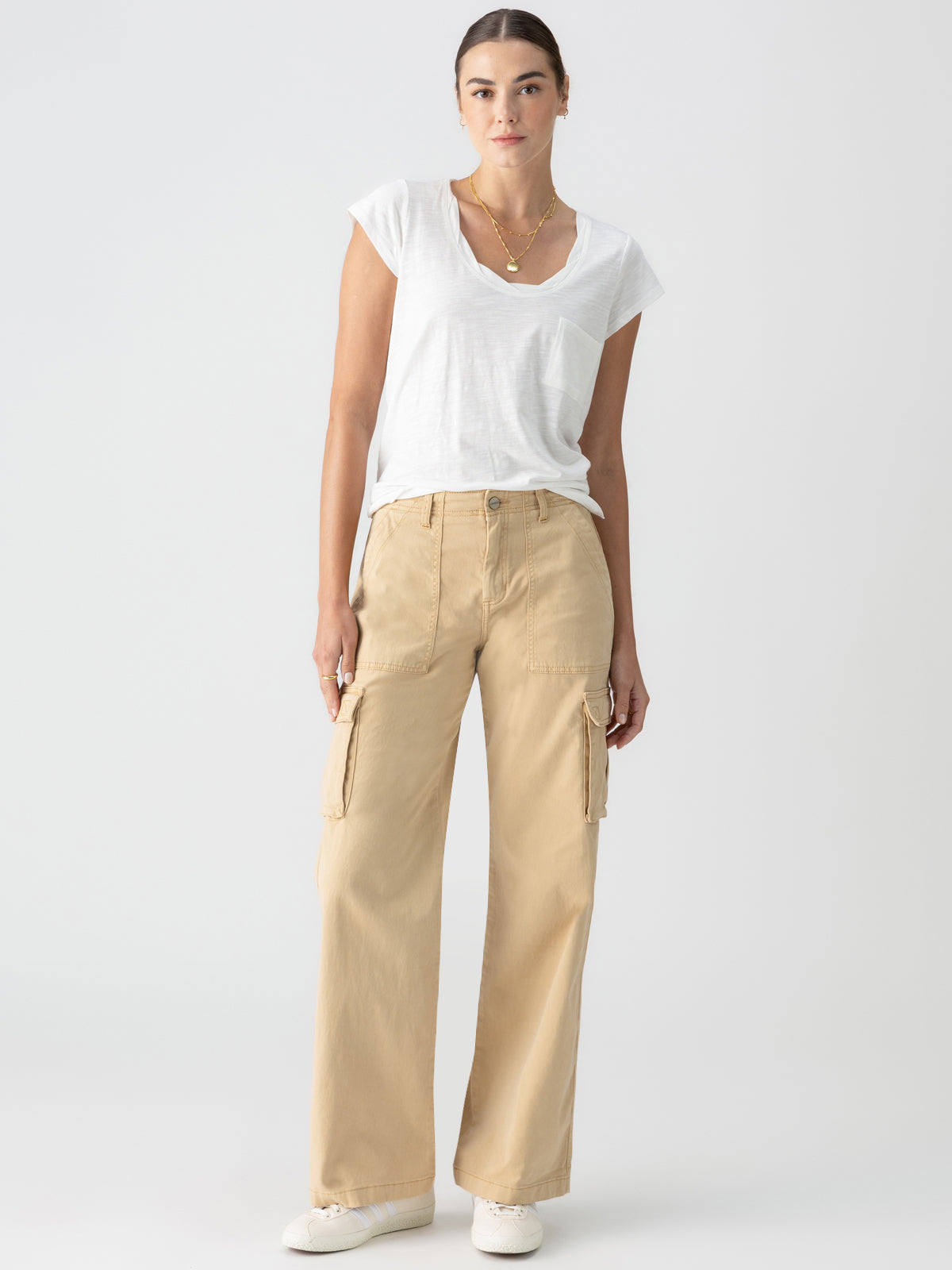 A woman stands against a plain background, wearing a white V-neck T-shirt and the **Reissue Cargo True Khaki** pants from **Sanctuary Clothing**, which feature large pockets. She completes her look with white sneakers, a simple necklace, and her hair pulled back.