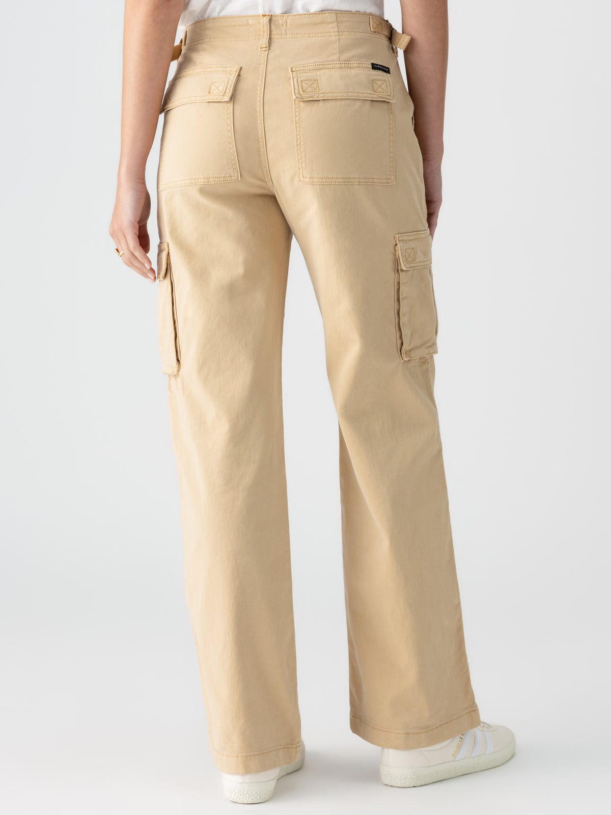 A person is shown from the back, wearing Sanctuary Clothing's Reissue Cargo True Khaki pants, featuring multiple pockets including side cargo pockets. They are also wearing a white shirt tucked into the pants and white shoes.