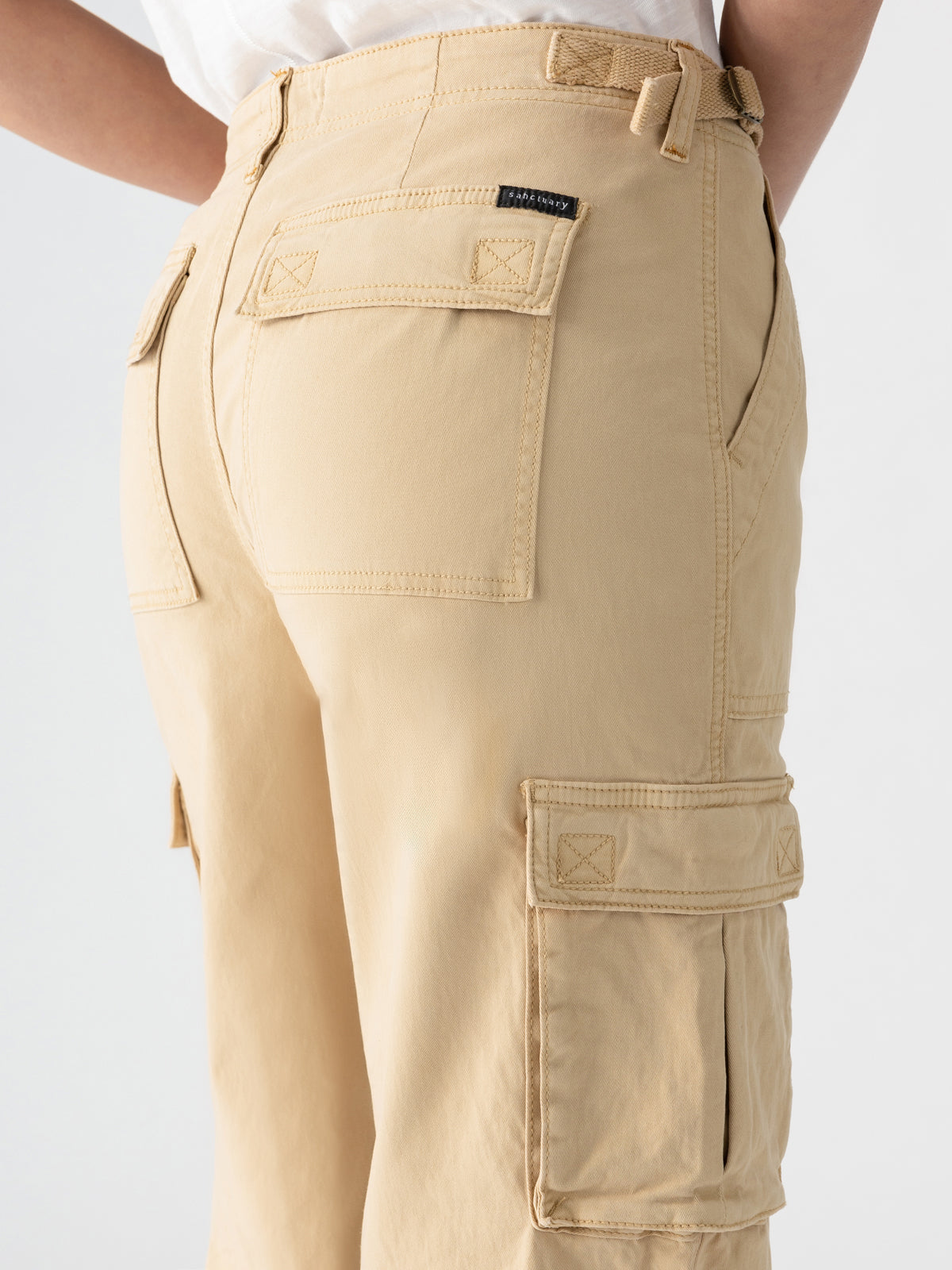 The image features a close-up of a person wearing the Reissue Cargo True Khaki pants by Sanctuary Clothing, showcasing their light brown hue, multiple pockets, belt loops, and intricate stitching details. The pants are paired with a white shirt and set against a plain white background.