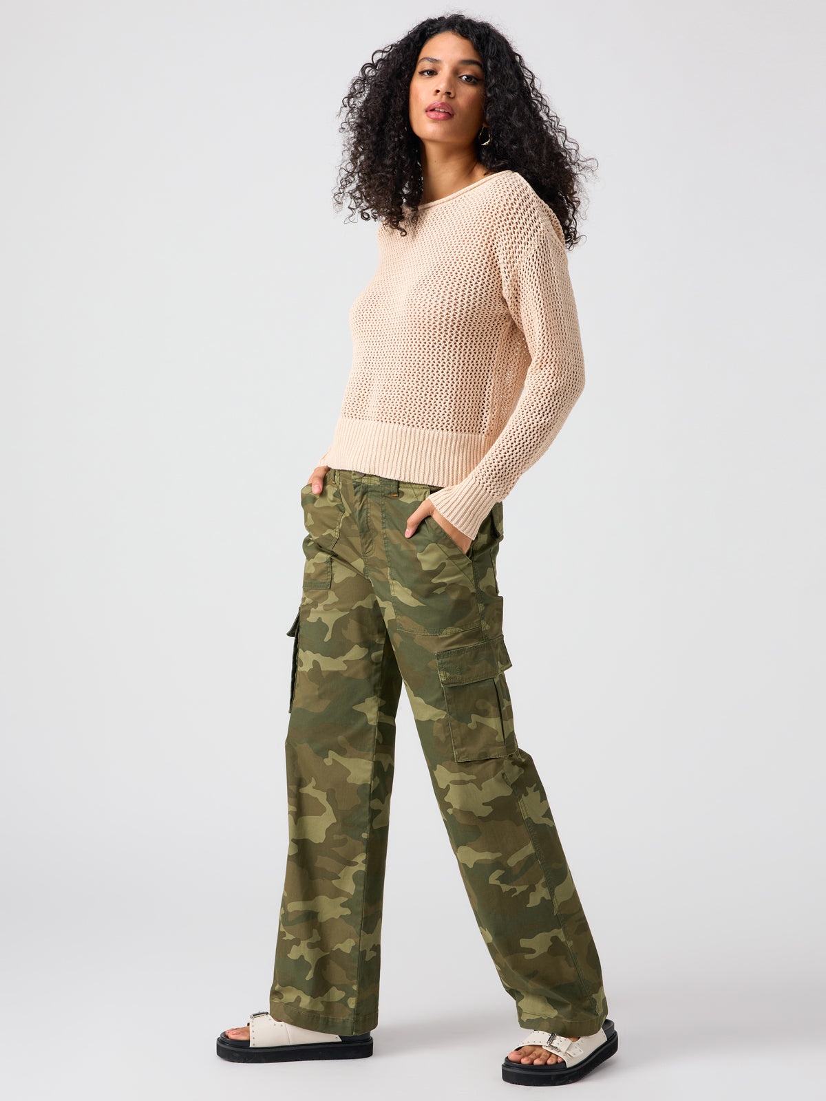 A person with long, curly hair is wearing a light beige, long-sleeved mesh sweater, Reissue Cargo Pant Renew Camo by Sanctuary Clothing, and white flat sandals. They are standing and facing slightly to the side against a plain background.