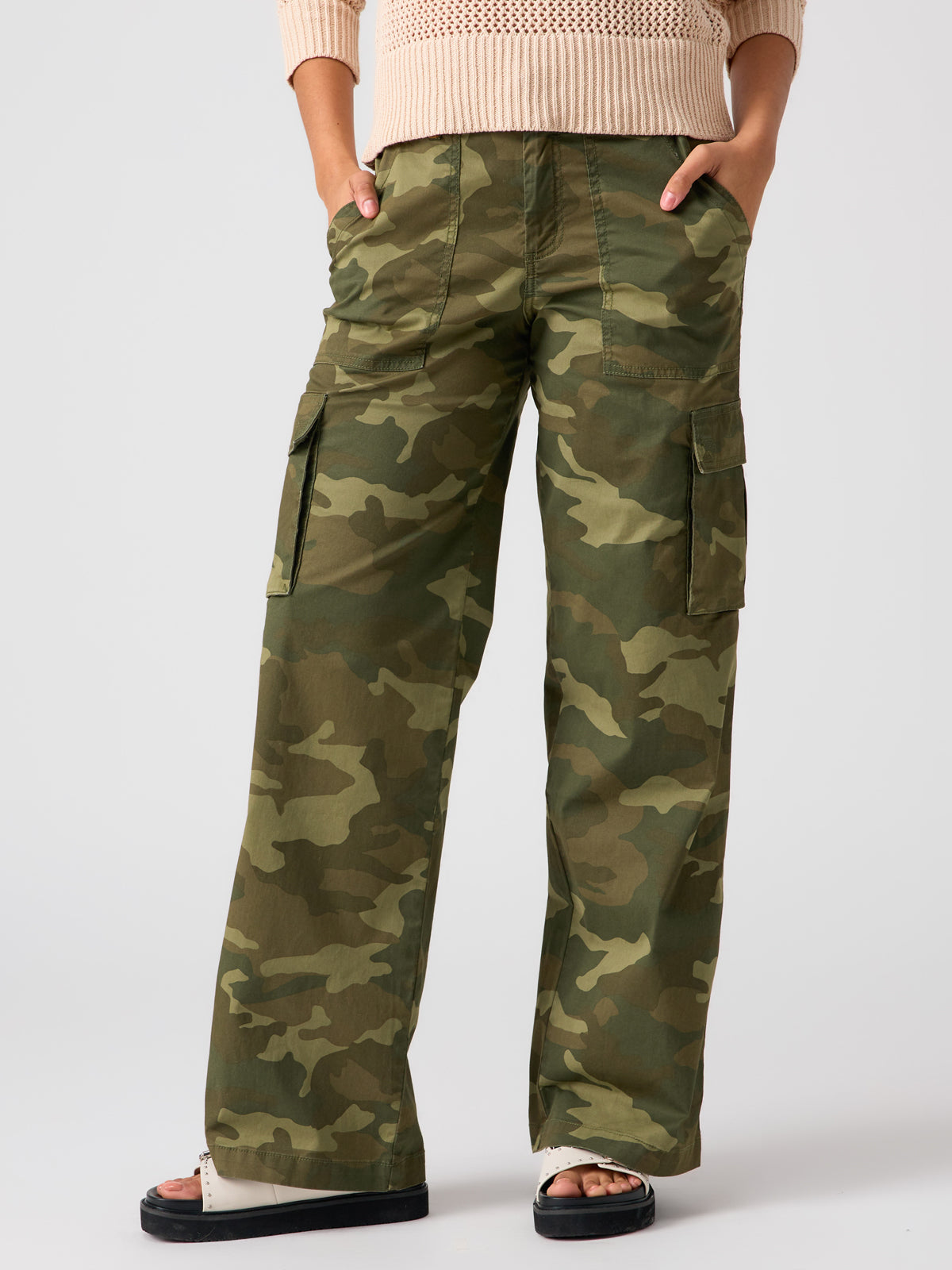 A person is wearing the Reissue Cargo Pant Renew Camo by Sanctuary Clothing with hands in the pockets. They are also wearing a beige sweater and black sandals with white soles. The background is plain white.