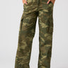 A person is wearing the Reissue Cargo Pant Renew Camo by Sanctuary Clothing with hands in the pockets. They are also wearing a beige sweater and black sandals with white soles. The background is plain white.