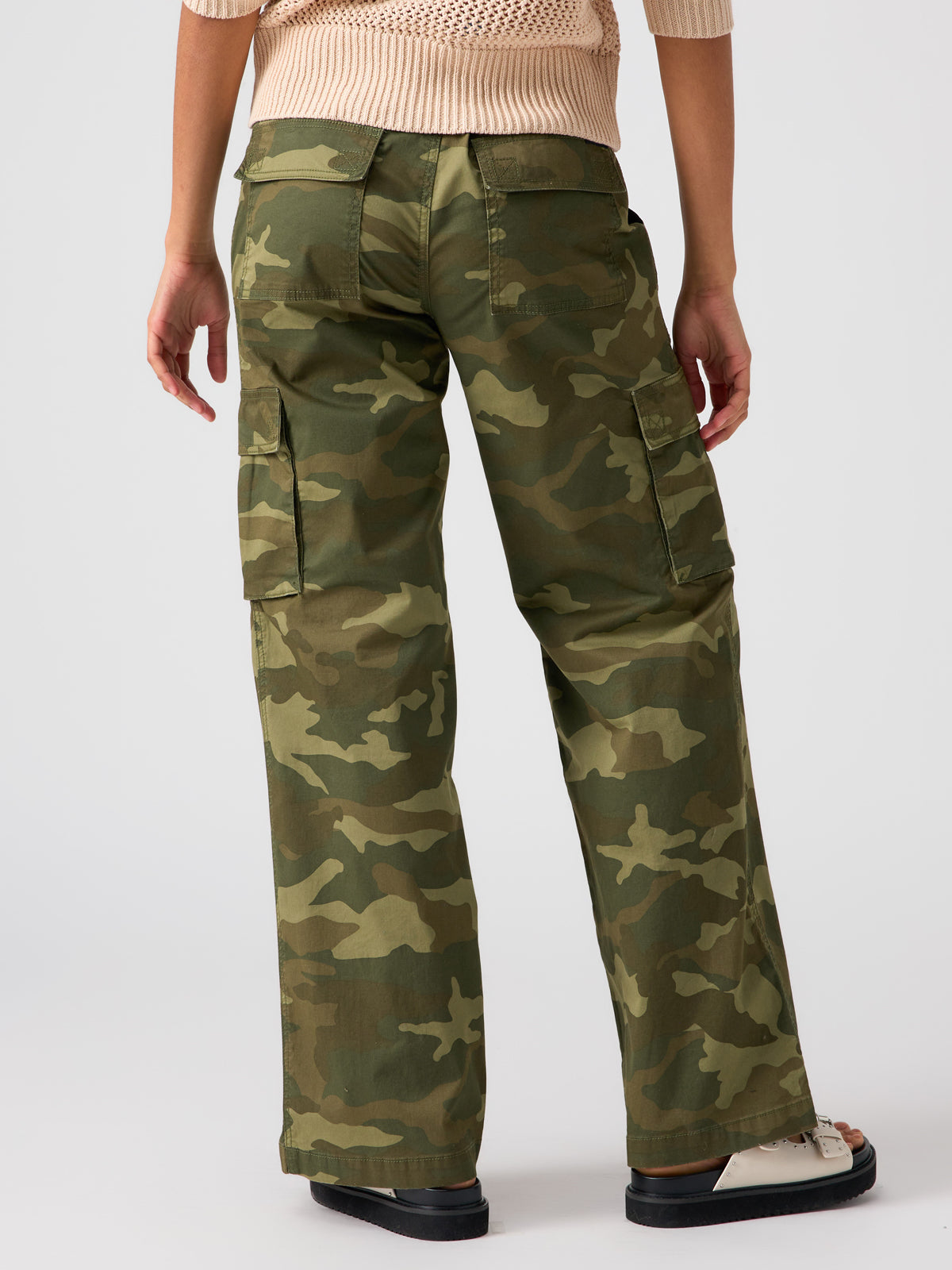 A person is standing with their back to the camera, wearing the Sanctuary Clothing Reissue Cargo Pant Renew Camo, which features green camouflage and large side pockets. They are also sporting a light-colored sweater and white shoes. The background is plain and white.