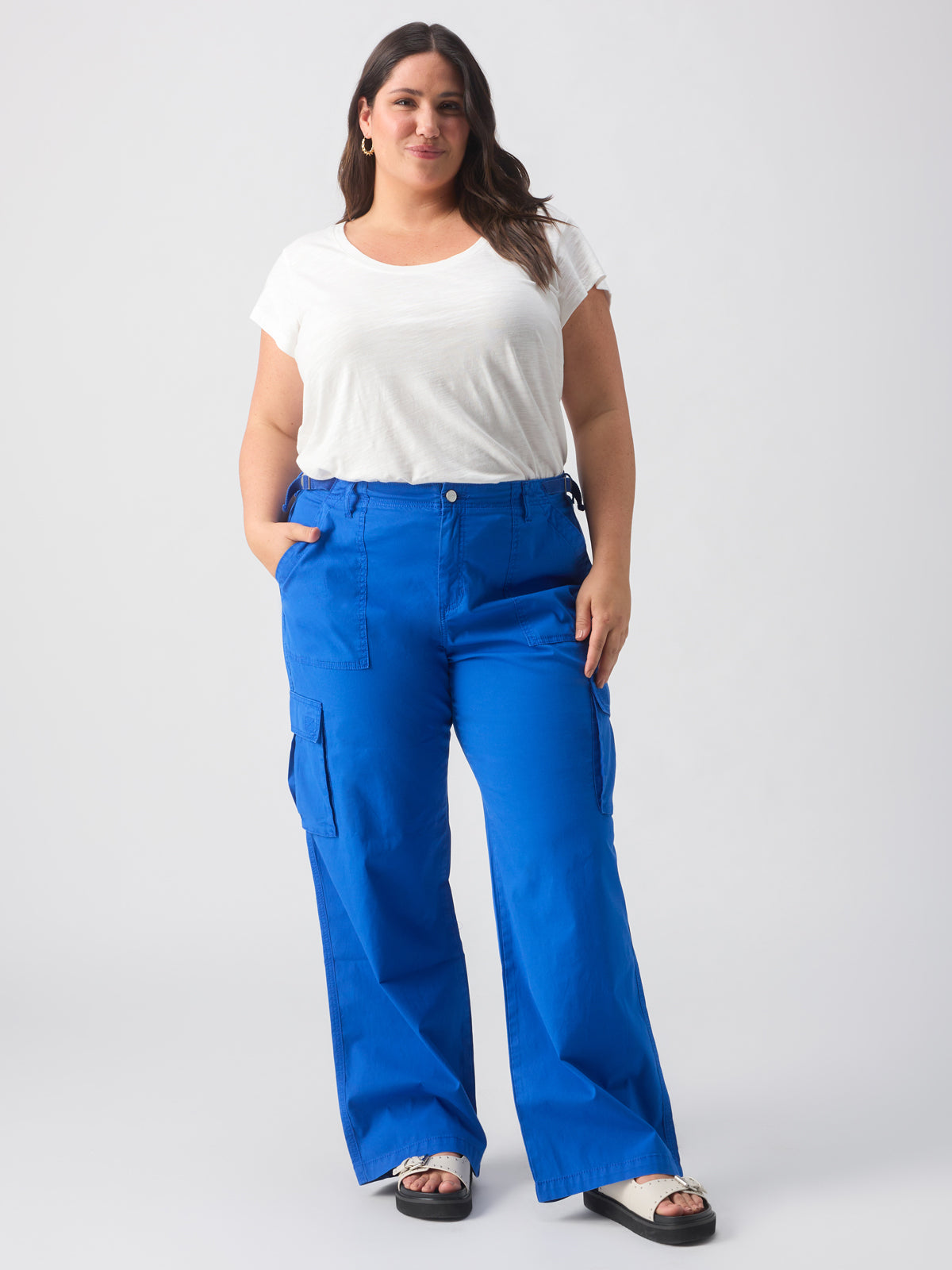 A woman stands against a plain white background, hands in the pockets of her bright blue Reissue Cargo Ocean Blue Inclusive Collection pants from Sanctuary Clothing. She is wearing a white t-shirt and beige sandals, with long brown hair styled loosely over her shoulders. She gazes confidently at the camera.