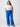 A woman stands against a plain white background, hands in the pockets of her bright blue Reissue Cargo Ocean Blue Inclusive Collection pants from Sanctuary Clothing. She is wearing a white t-shirt and beige sandals, with long brown hair styled loosely over her shoulders. She gazes confidently at the camera.